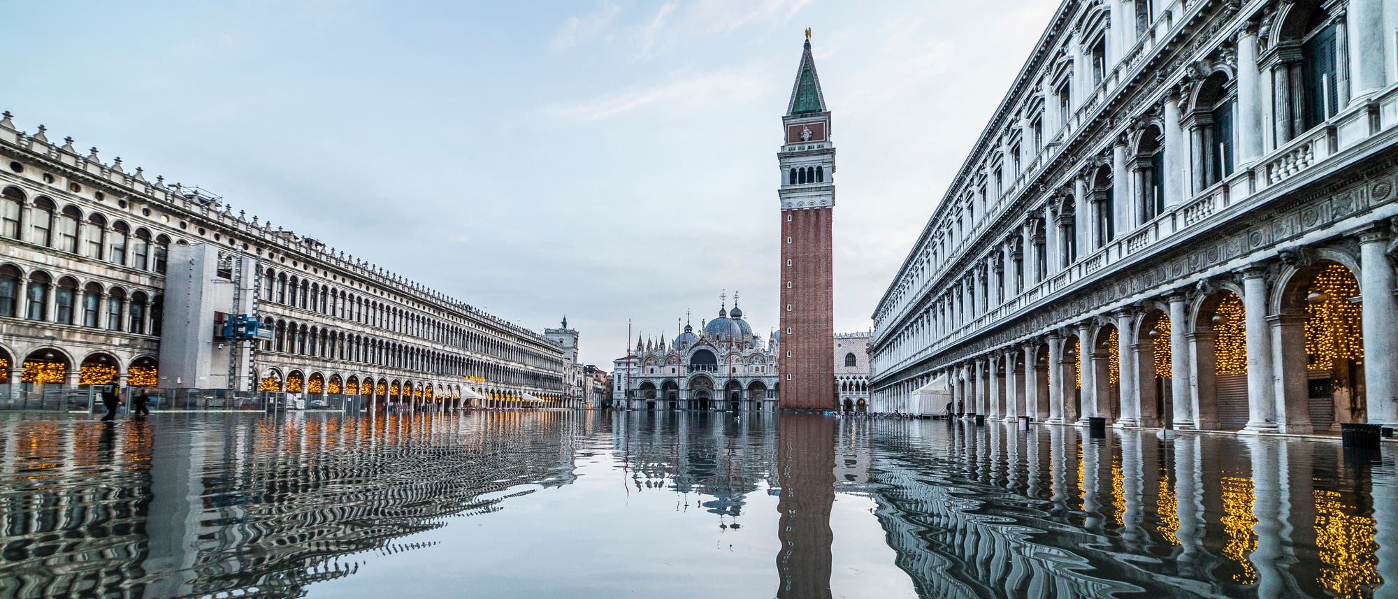 Eine &uuml;berflutete Piazza mit historischen Geb&auml;uden und einem hohen Glockenturm im Hintergrund. Die Wasseroberfl&auml;che spiegelt die Architektur wider, w&auml;hrend die Lichter in den Arkaden auf der rechten Seite leuchten. Der Himmel ist bew&ouml;lkt, was eine ruhige und reflektierende Atmosph&auml;re schafft.