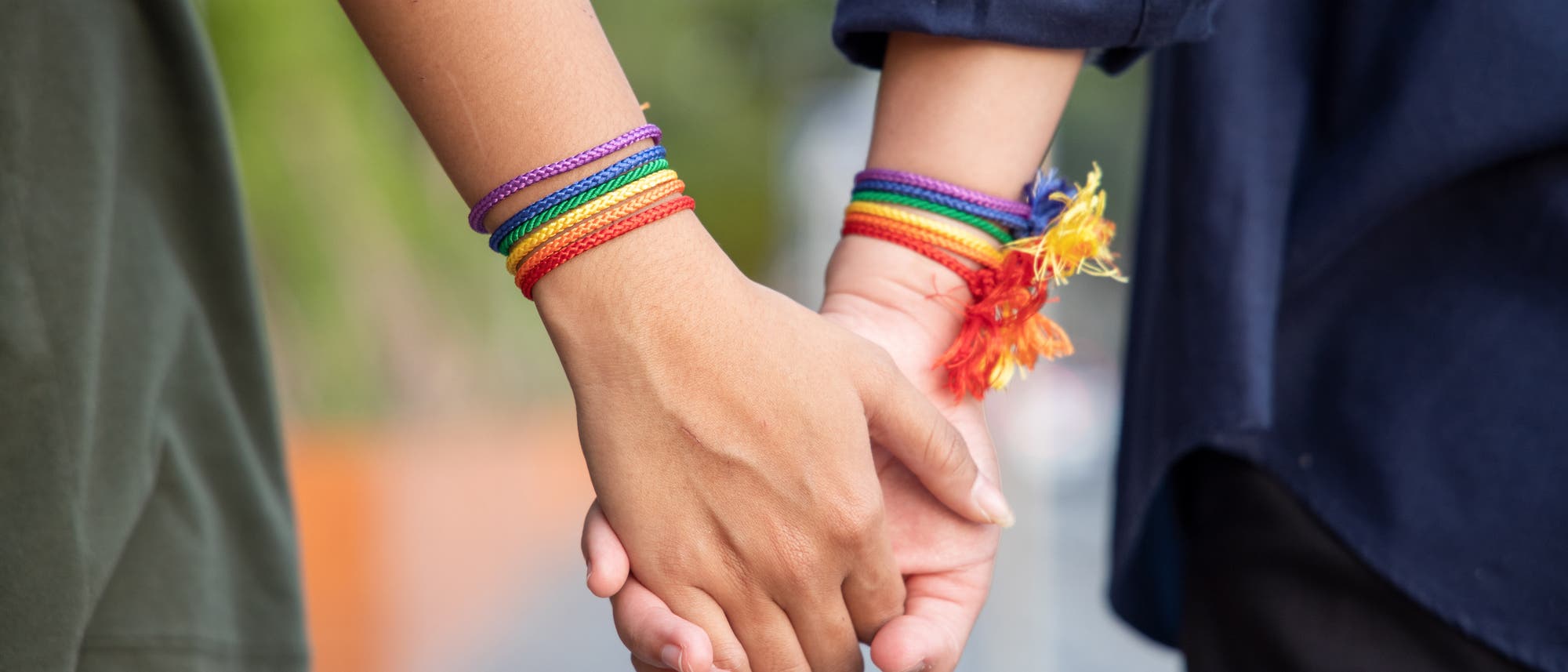 Zwei Personen halten sich an den Händen, beide tragen Armbänder in Regenbogenfarben, die ein Symbol für die LGBTQ-Community sind. Die Szene vermittelt ein Gefühl von Zusammengehörigkeit und Unterstützung. Der Hintergrund ist unscharf, was den Fokus auf die Hände und Armbänder lenkt. Zwei Personen halten sich an den Händen, beide tragen Armbänder in Regenbogenfarben, die ein Symbol für die LGBTQ-Community sind. Die Szene vermittelt ein Gefühl von Zusammengehörigkeit und Unterstützung. Der Hintergrund ist unscharf, was den Fokus auf die Hände und Armbänder lenkt.