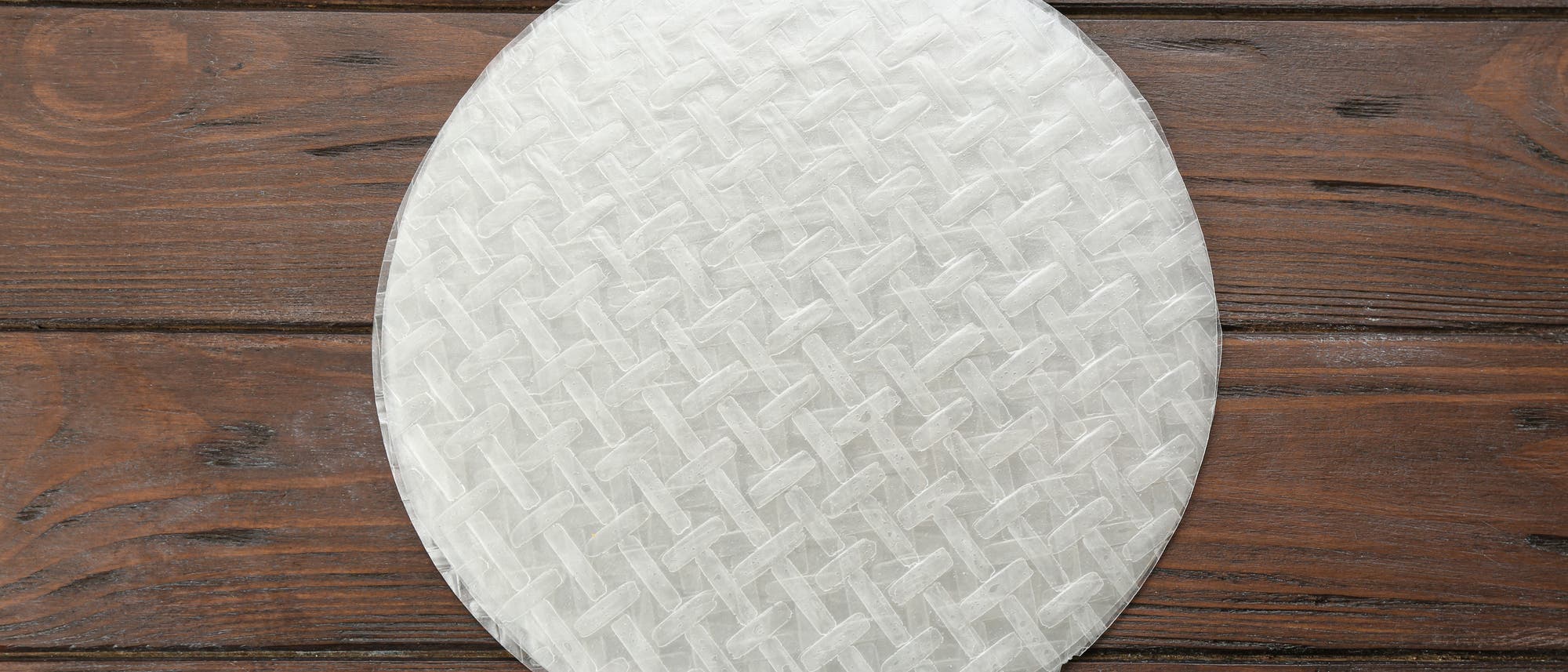 Ein einzelnes rundes Reispapierblatt liegt auf einer dunklen Holzoberfläche. Das Reispapier hat eine durchscheinende, gewebte Textur. Es wird häufig in der asiatischen Küche verwendet, um Frühlingsrollen oder Sommerrollen herzustellen.