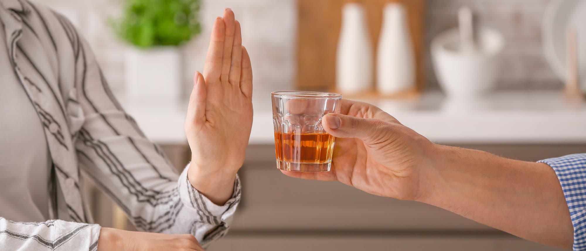 Eine Person lehnt ein Glas mit einem alkoholischen Getr&auml;nk ab, indem sie die Hand in einer abwehrenden Geste hebt. Die Szene spielt sich in einer h&auml;uslichen Umgebung ab, mit unscharfem Hintergrund, der K&uuml;chenutensilien und Pflanzen zeigt. Das Bild vermittelt eine Botschaft der Abstinenz oder Ablehnung von Alkohol.