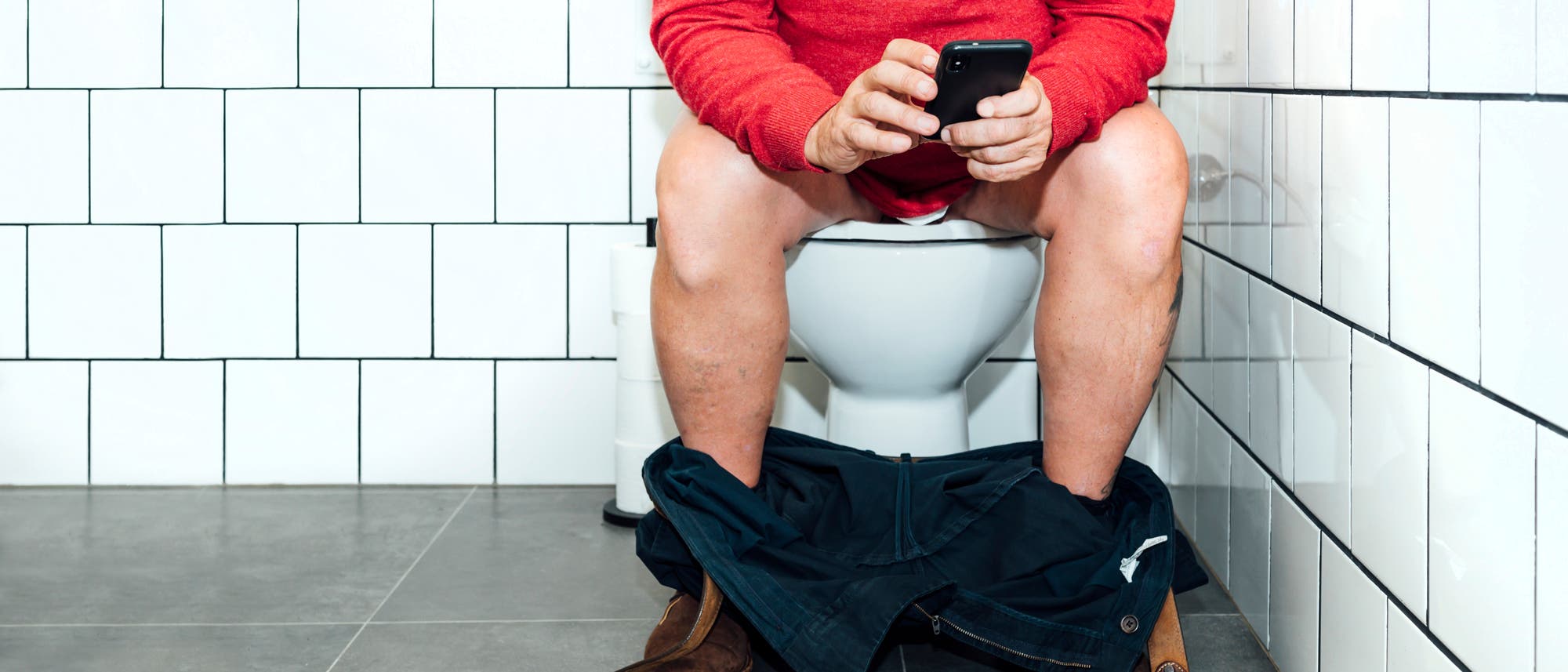 Eine Person in einem roten Pullover sitzt auf einer Toilette in einem gefliesten Badezimmer und hält ein Smartphone in den Händen. Die Hose ist heruntergelassen, und die Person scheint auf das Telefon zu schauen.