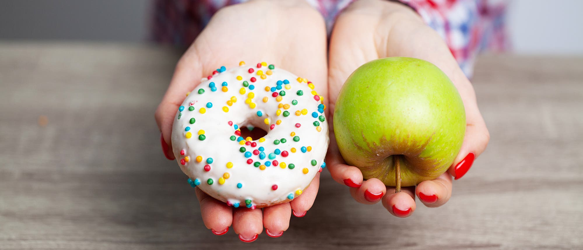 Eine Person hält in der linken Hand einen Donut mit weißer Glasur und bunten Streuseln und in der rechten Hand einen grünen Apfel. Die Hände sind vor einem Holztisch ausgestreckt. Die Person trägt ein kariertes Hemd. Das Bild thematisiert den Kontrast zwischen ungesunder und gesunder Ernährung. Eine Person hält in der linken Hand einen Donut mit weißer Glasur und bunten Streuseln und in der rechten Hand einen grünen Apfel. Die Hände sind vor einem Holztisch ausgestreckt. Die Person trägt ein kariertes Hemd. Das Bild thematisiert den Kontrast zwischen ungesunder und gesunder Ernährung.