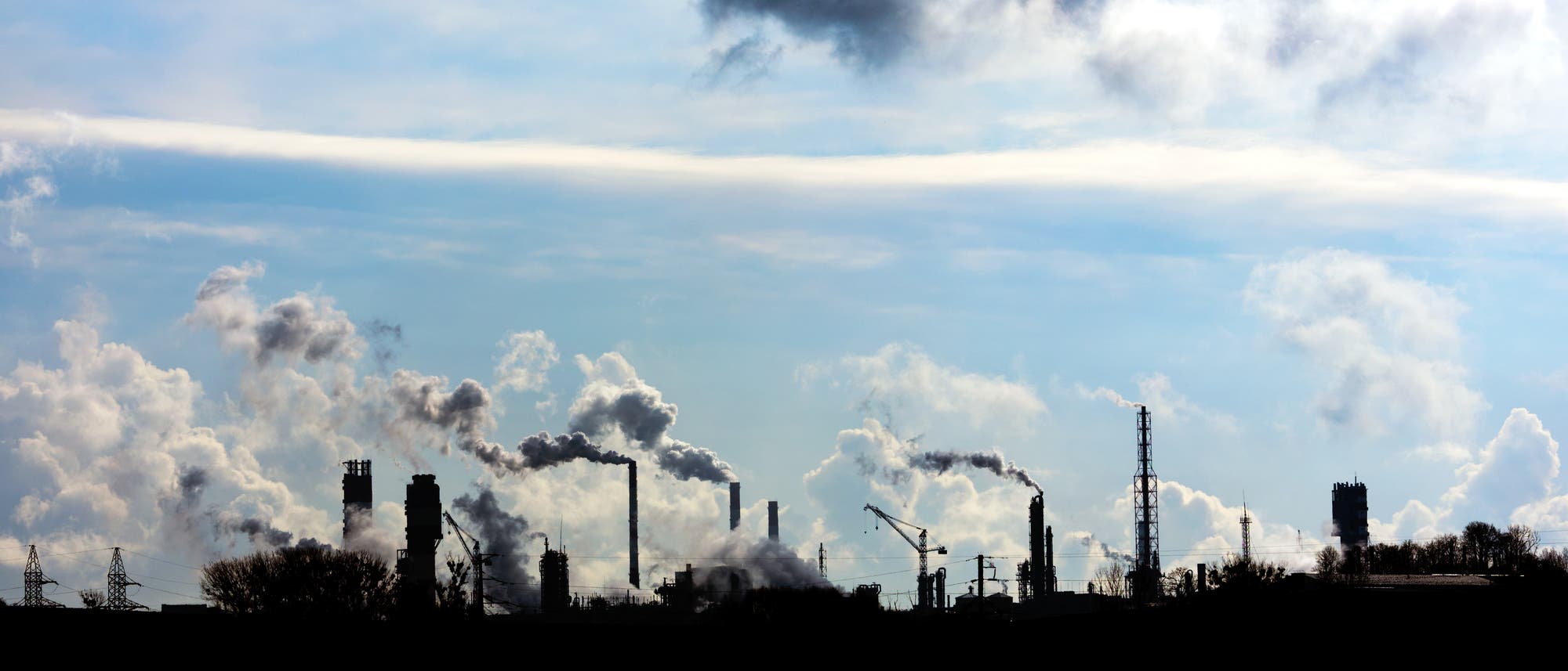 Industrieanlage mit mehreren Schornsteinen, die Rauch in den Himmel abgeben. Der Himmel ist bewölkt, und die Silhouetten der Fabrikstrukturen sind vor dem Hintergrund des Himmels sichtbar. Die Szene vermittelt ein Gefühl von industrieller Aktivität und Umweltbelastung.