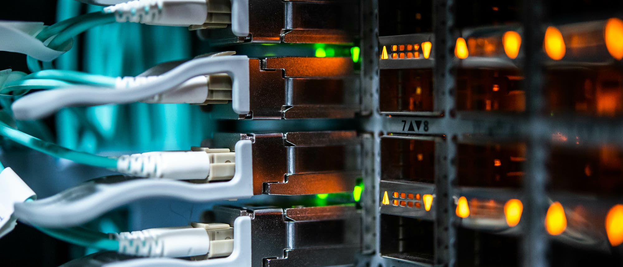 Nahaufnahme eines Serverracks mit mehreren eingesteckten Netzwerkkabeln. Die Kabel sind mit leuchtenden gr&uuml;nen und orangefarbenen LED-Anzeigen verbunden, die den Netzwerkstatus anzeigen. Die Umgebung ist in einem ged&auml;mpften, bl&auml;ulichen Licht gehalten, das die technische Atmosph&auml;re unterstreicht.