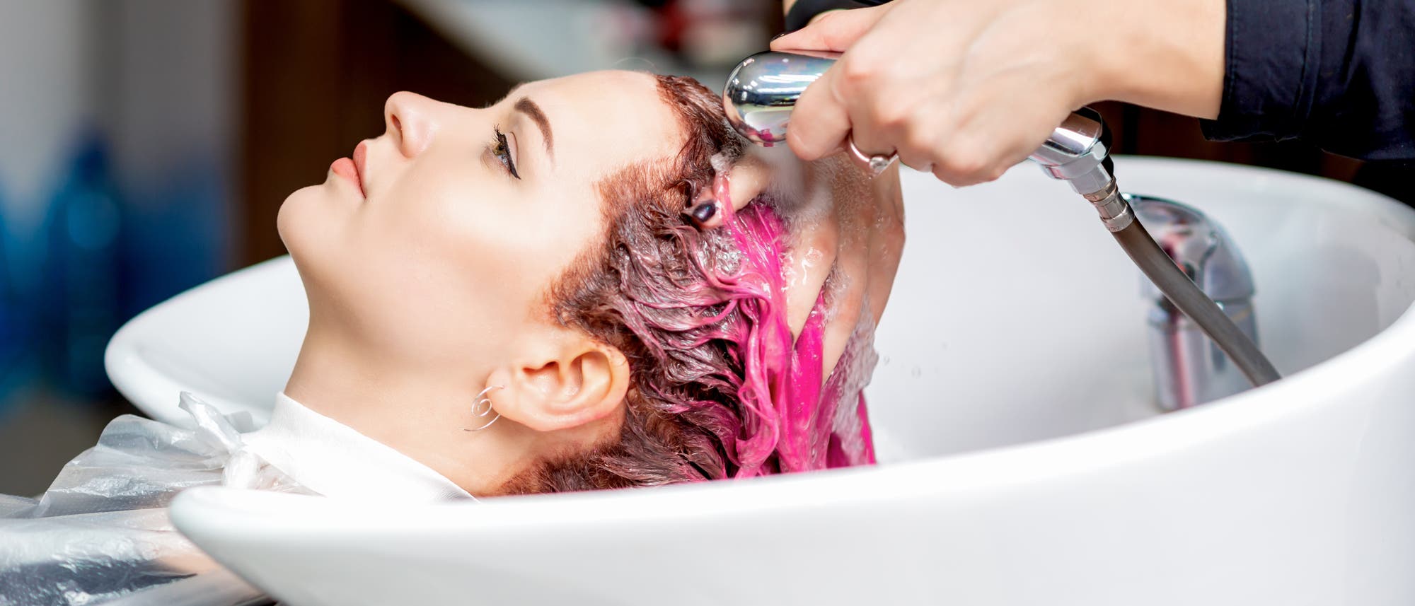 Eine Person liegt in einem Friseursalon an einem Waschbecken, während eine andere Person ihr die Haare wäscht. Die Haare sind mit einer pinken Farbe behandelt. Die Szene zeigt den Prozess des Haarewaschens mit einem Duschkopf. Im Hintergrund sind unscharfe Details des Salons zu sehen. Eine Person liegt in einem Friseursalon an einem Waschbecken, während eine andere Person ihr die Haare wäscht. Die Haare sind mit einer pinken Farbe behandelt. Die Szene zeigt den Prozess des Haarewaschens mit einem Duschkopf. Im Hintergrund sind unscharfe Details des Salons zu sehen.