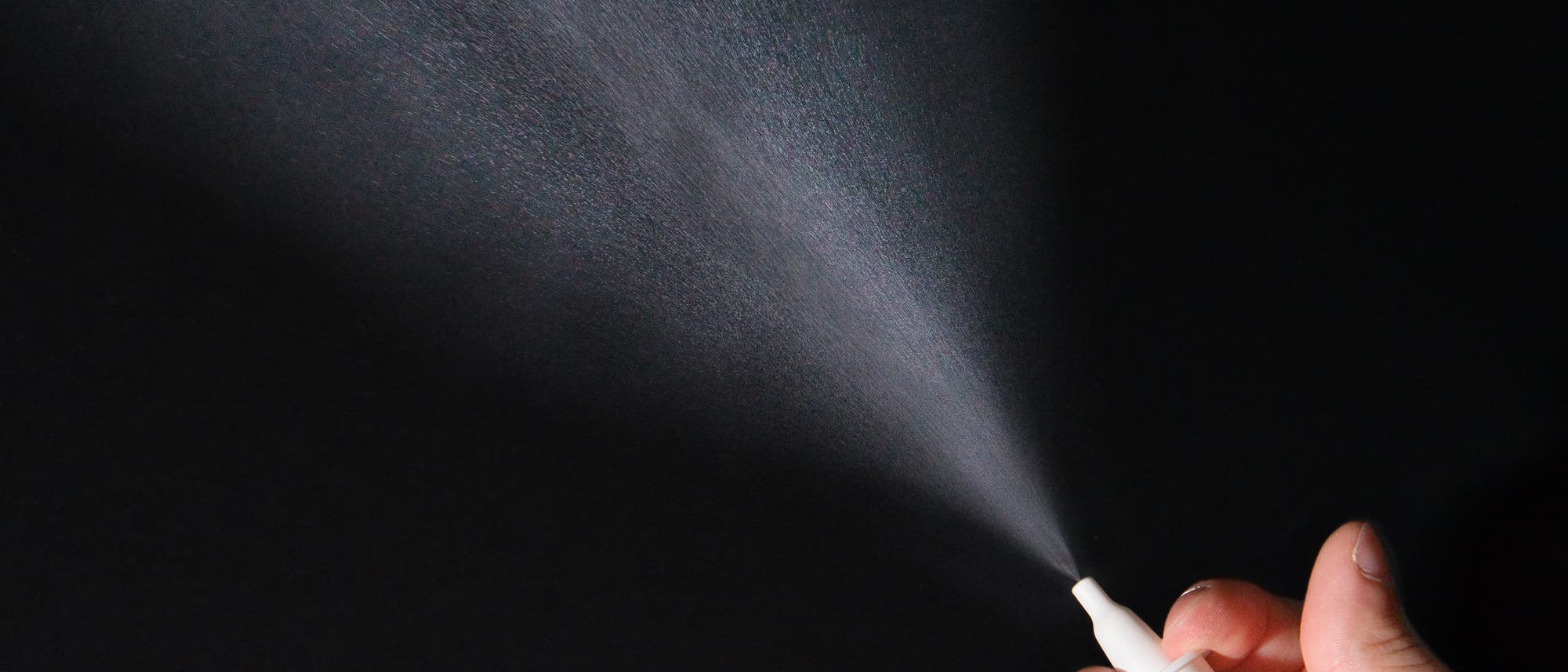 Finger pumpen Nasenspray in die Luft, das entstehende Aerosol ist vor schwarzem Hintergrund gut sichtbar.