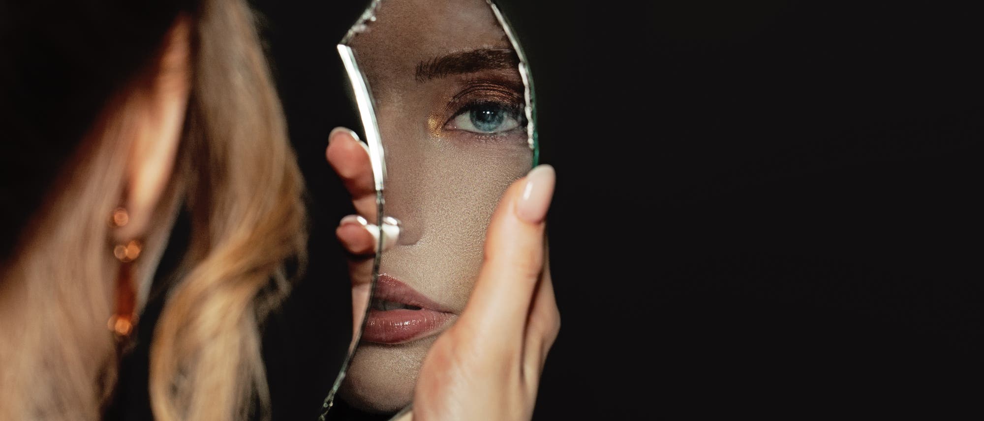 Blonde Frau schaut vor dunklem Hintergrund in Scherbe von zerbrochenen Spiegel.