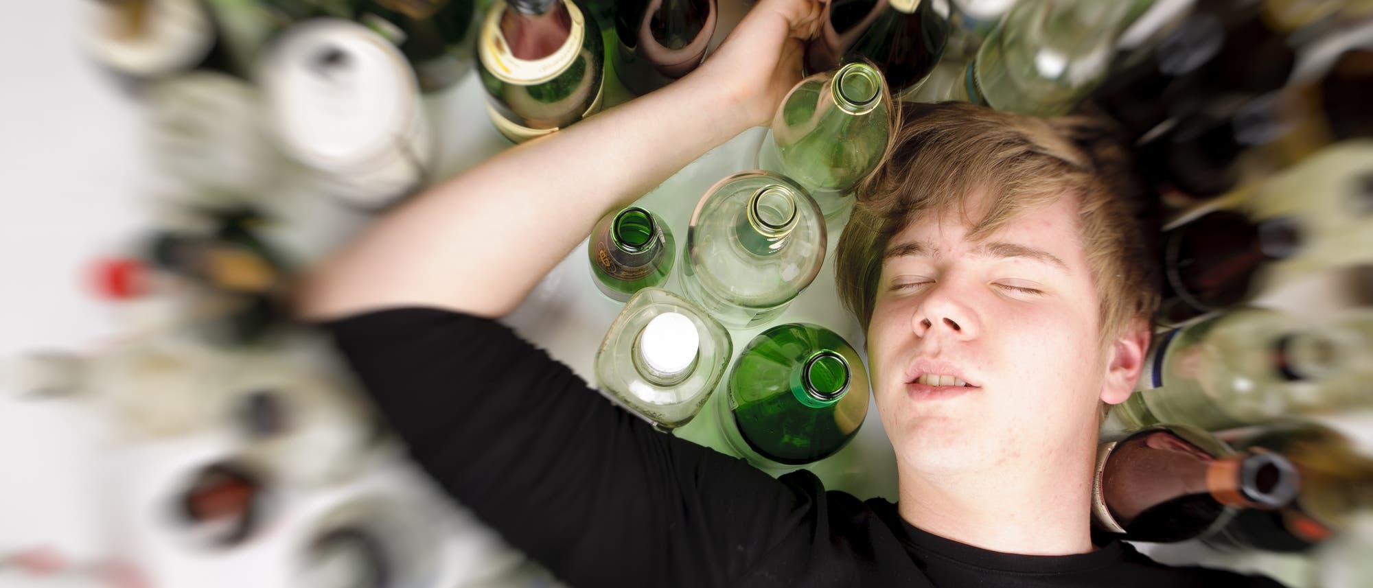 Ein männlicher Jugendlicher liegt im Rausch schlafend neben vielen leeren Flaschen auf dem Boden.
