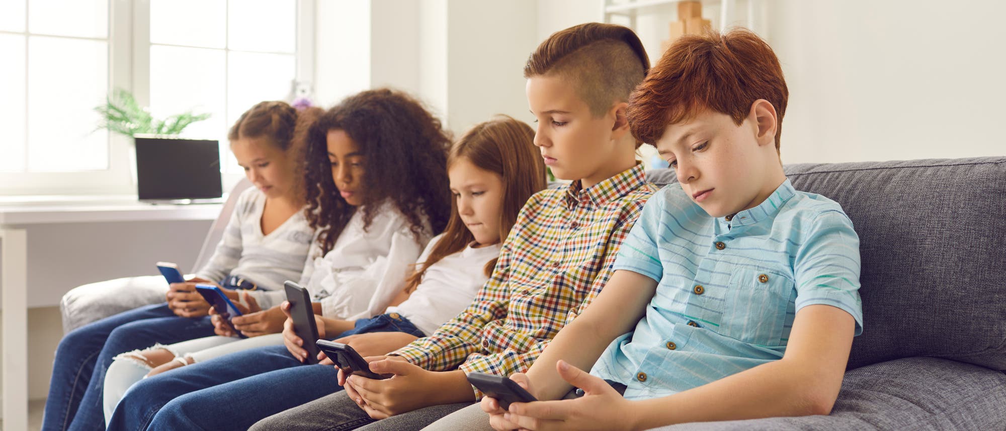 Eine Gruppe von fünf Kindern sitzt nebeneinander auf einem Sofa in einem hellen Raum. Jedes Kind hält ein Smartphone in der Hand und ist darauf konzentriert. Im Hintergrund sind ein Fenster und ein Regal mit einer Pflanze zu sehen. Die Szene vermittelt den Eindruck von moderner Technologie und digitaler Interaktion.