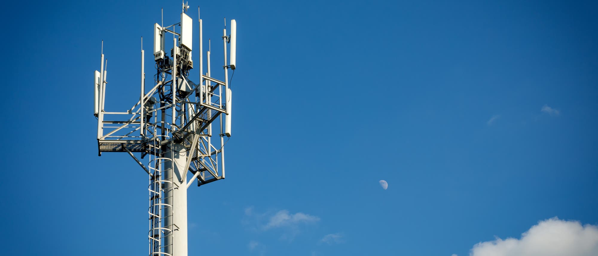 Ein Mobilfunkmast mit mehreren Antennen ragt in den klaren blauen Himmel. Im Hintergrund ist der Mond sichtbar, umgeben von wenigen kleinen Wolken. Der Mast symbolisiert moderne Kommunikationstechnologie.