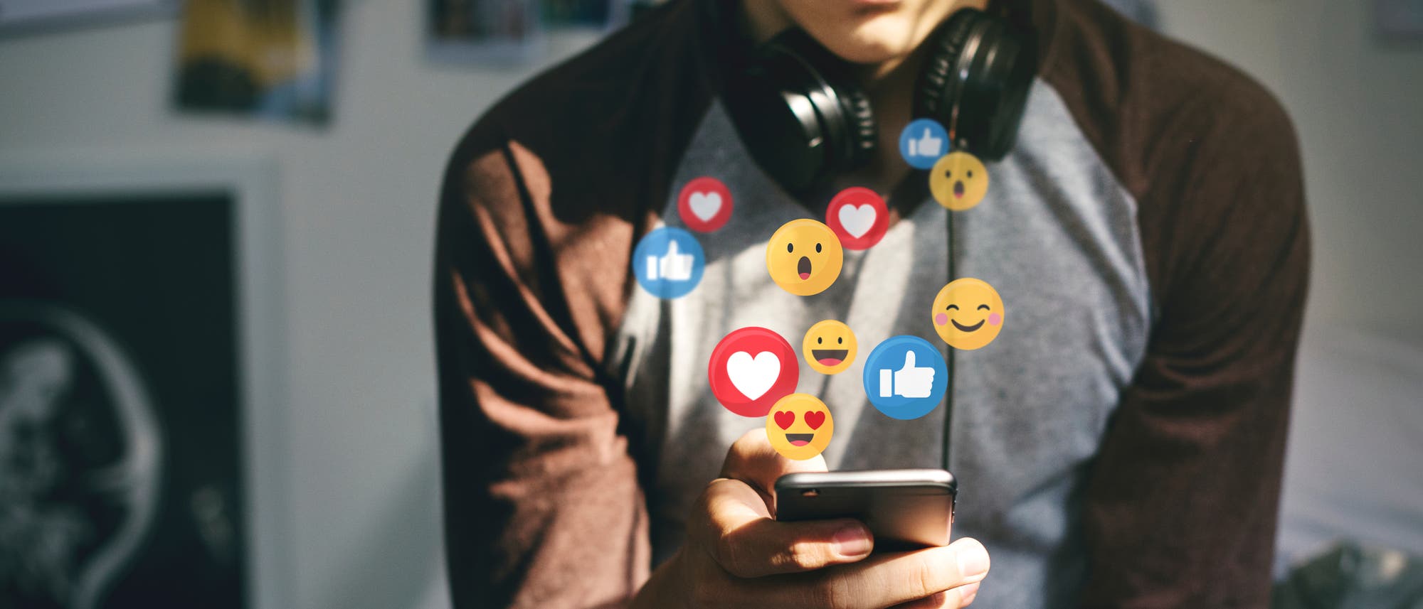 Ein Jugendlicher h&auml;lt ein Smartphone in der Hand. Um ihn herum sind verschiedenen Social-Media-Emojis eingeblendet, darunter Herzen, Daumen hoch und lachende Gesichter. 