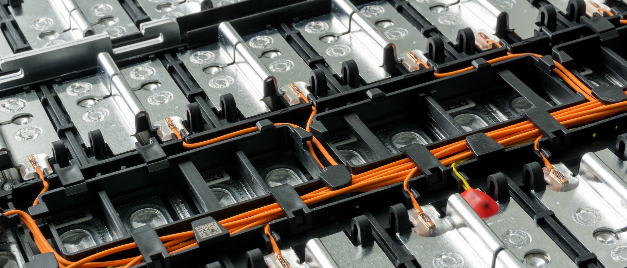 Nahaufnahme einer Batterieeinheit mit mehreren Zellen, verbunden durch orangefarbene Kabel und Metallverbindungen. Die Struktur zeigt eine Anordnung von rechteckigen Modulen, die in einem Rastermuster angeordnet sind. Die Oberfläche der Zellen ist metallisch glänzend, und die Kabel sind sorgfältig in schwarzen Halterungen fixiert. Keine sichtbaren Texte oder URLs. Nahaufnahme einer Batterieeinheit mit mehreren Zellen, verbunden durch orangefarbene Kabel und Metallverbindungen. Die Struktur zeigt eine Anordnung von rechteckigen Modulen, die in einem Rastermuster angeordnet sind. Die Oberfläche der Zellen ist metallisch glänzend, und die Kabel sind sorgfältig in schwarzen Halterungen fixiert. Keine sichtbaren Texte oder URLs.