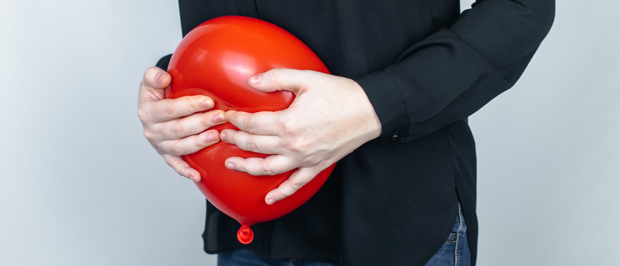 Eine Person in einem schwarzen Hemd h&auml;lt einen roten Ballon vor ihrem Bauch. Der Hintergrund ist neutral und hell. Der Fokus liegt auf den H&auml;nden, die den Ballon umschlie&szlig;en.