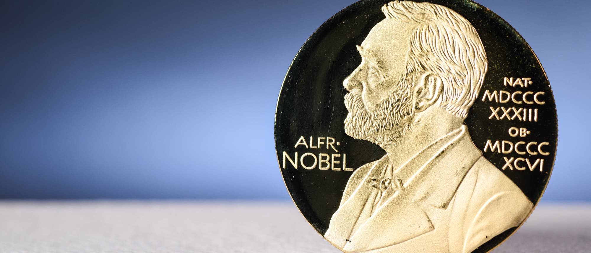 Eine goldene Medaille mit dem Profil eines Mannes, der nach rechts schaut, auf einem unscharfen blauen Hintergrund. Auf der Medaille steht &bdquo;ALFR. NOBEL&ldquo; sowie r&ouml;mische Zahlen. Die Medaille liegt auf einer hellen Oberfl&auml;che.