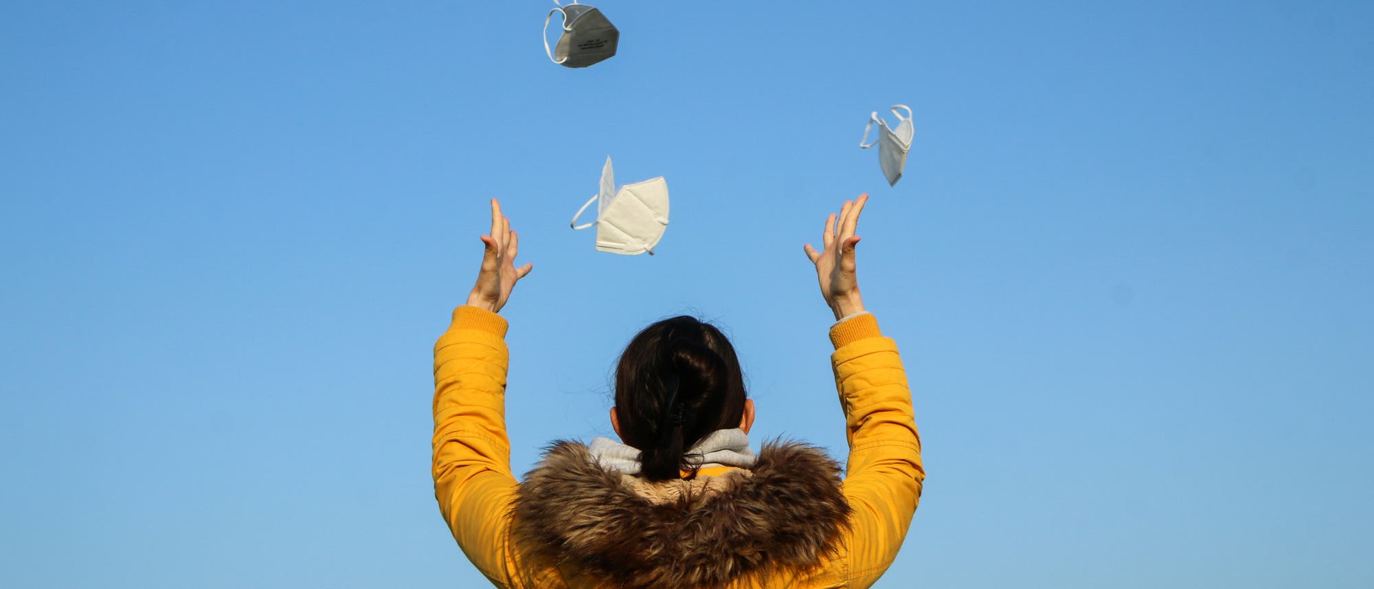 Eine Frau von hinten in gelber Winterjacke, die vor einem blauen Himmel mehrere FFP2-Masken in die Luft wirft.