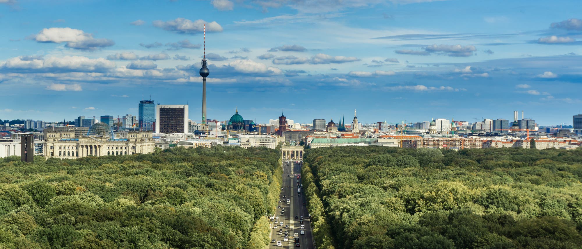 Blick auf die Skyline von Berlin mit dem markanten Fernsehturm und dem Brandenburger Tor im Zentrum. Im Vordergrund erstreckt sich der Tiergarten, ein großer, grüner Park, durch den eine Straße mit fahrenden Autos führt. Die Stadtlandschaft dahinter zeigt eine Mischung aus historischen und modernen Gebäuden. Der Himmel ist blau mit vereinzelten Wolken.
