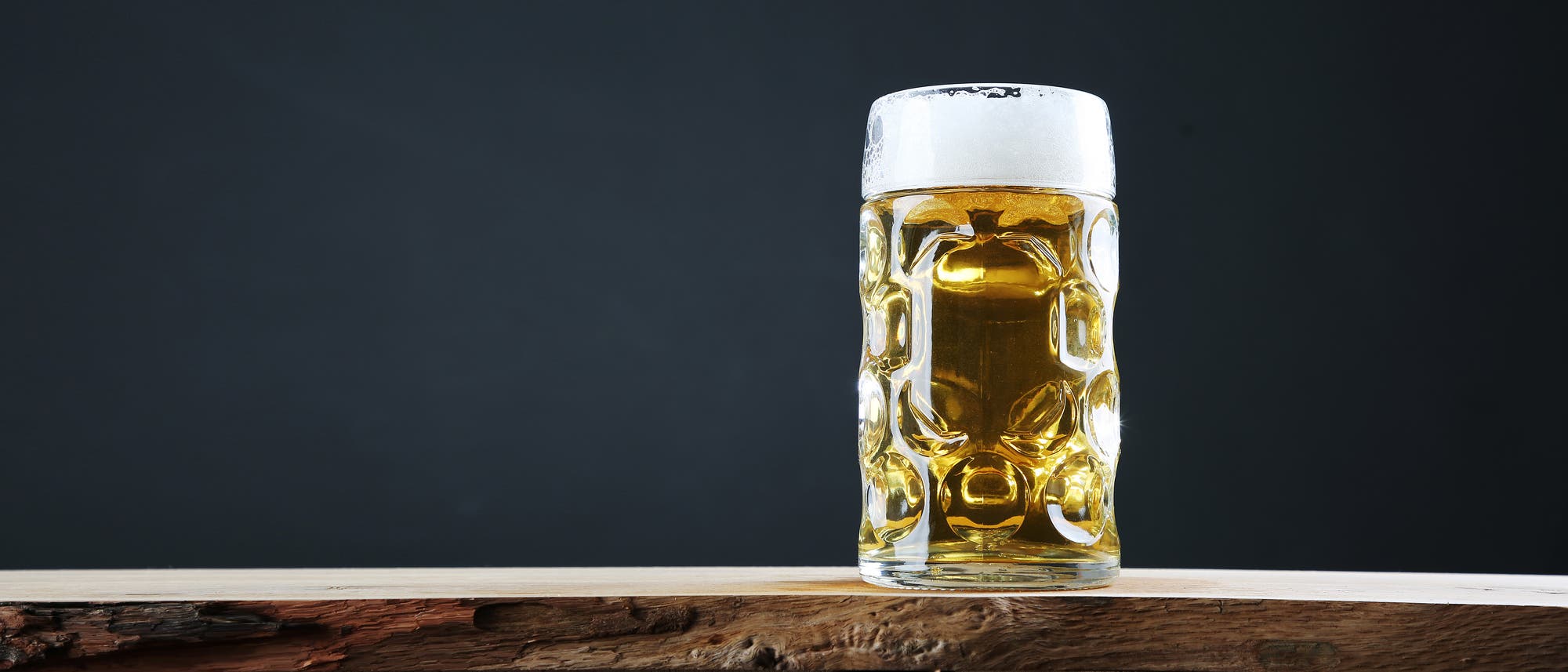 Ein großes, mit goldenem Bier gefülltes Glas mit Schaumkrone steht auf einem rustikalen Holztisch vor einem dunklen Hintergrund. Das Glas hat ein markantes, strukturiertes Design, das das Licht reflektiert.