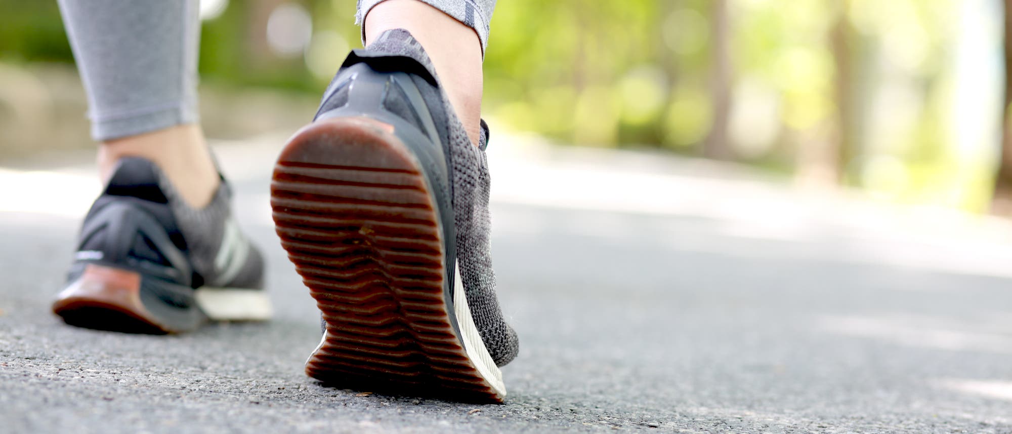 Eine Person in grauen Leggings geht auf einem asphaltierten Weg. Der Fokus liegt auf den Sohlen der schwarzen Sportschuhe. Im Hintergrund sind unscharf gr&uuml;ne B&auml;ume zu sehen, die auf eine nat&uuml;rliche Umgebung hinweisen.