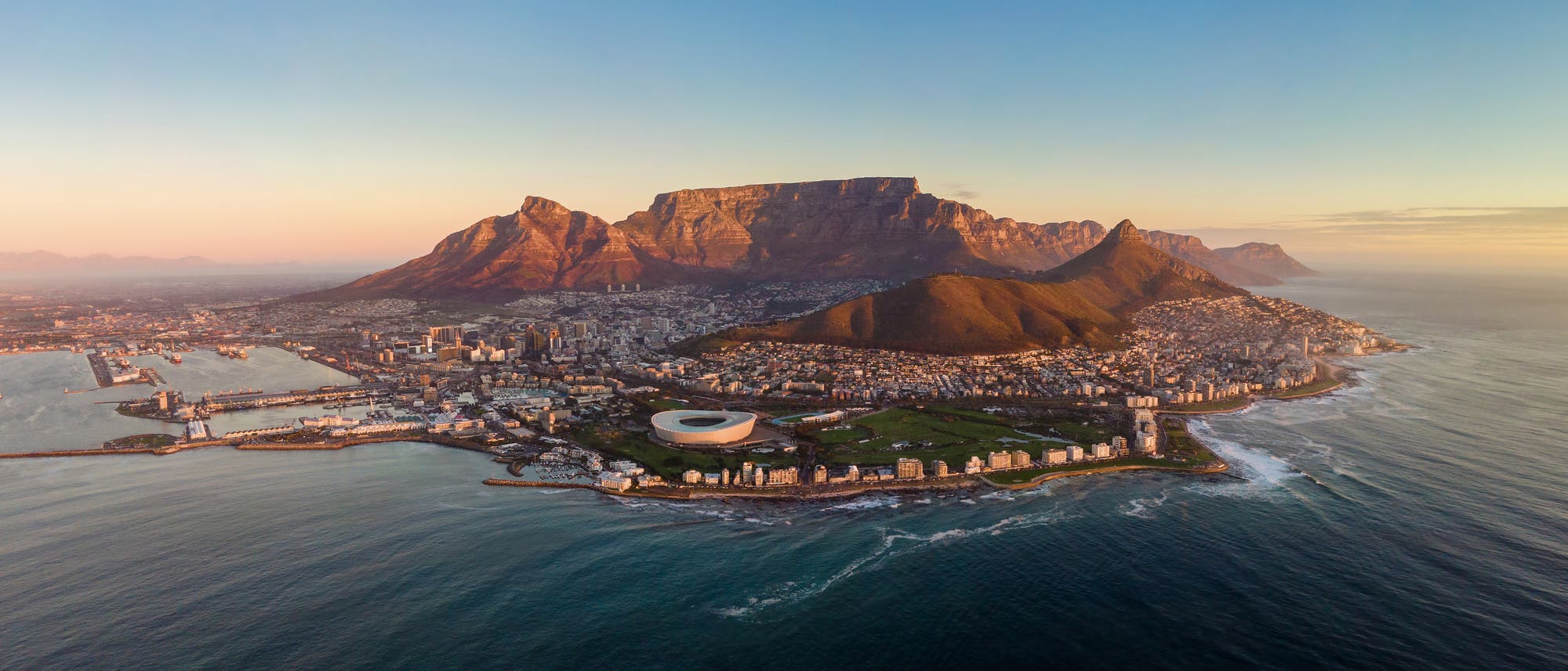 Luftaufnahme von Kapstadt, Südafrika, bei Sonnenuntergang. Im Vordergrund ist das markante Stadion zu sehen, umgeben von der Stadtlandschaft. Im Hintergrund erhebt sich der Tafelberg majestätisch. Die Küstenlinie umrahmt die Stadt, während das Meer ruhig wirkt. Keine Menschen sind sichtbar. Luftaufnahme von Kapstadt, Südafrika, bei Sonnenuntergang. Im Vordergrund ist das markante Stadion zu sehen, umgeben von der Stadtlandschaft. Im Hintergrund erhebt sich der Tafelberg majestätisch. Die Küstenlinie umrahmt die Stadt, während das Meer ruhig wirkt. Keine Menschen sind sichtbar.