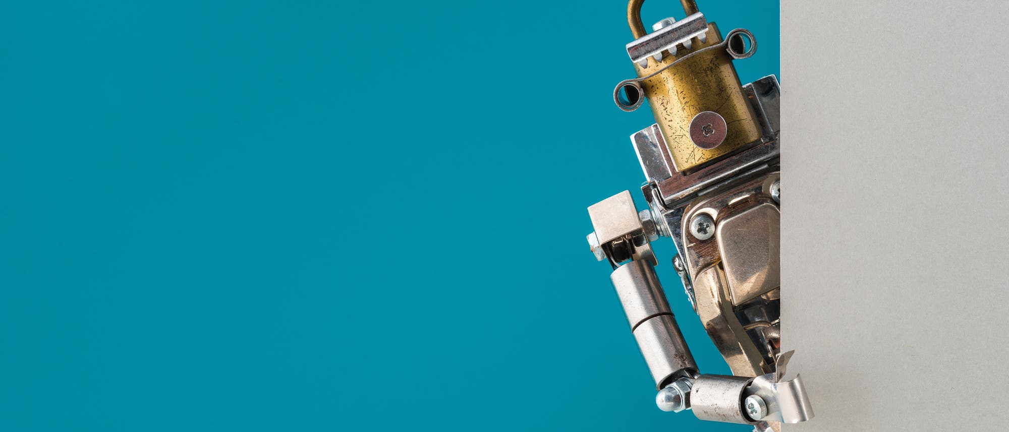 Ein kleiner Roboter aus Metallteilen schaut neugierig hinter einer grauen Wand hervor. Der Hintergrund ist einfarbig t&uuml;rkis. Der Roboter hat einen zylinderf&ouml;rmigen Kopf mit Schrauben und Muttern als Details.
