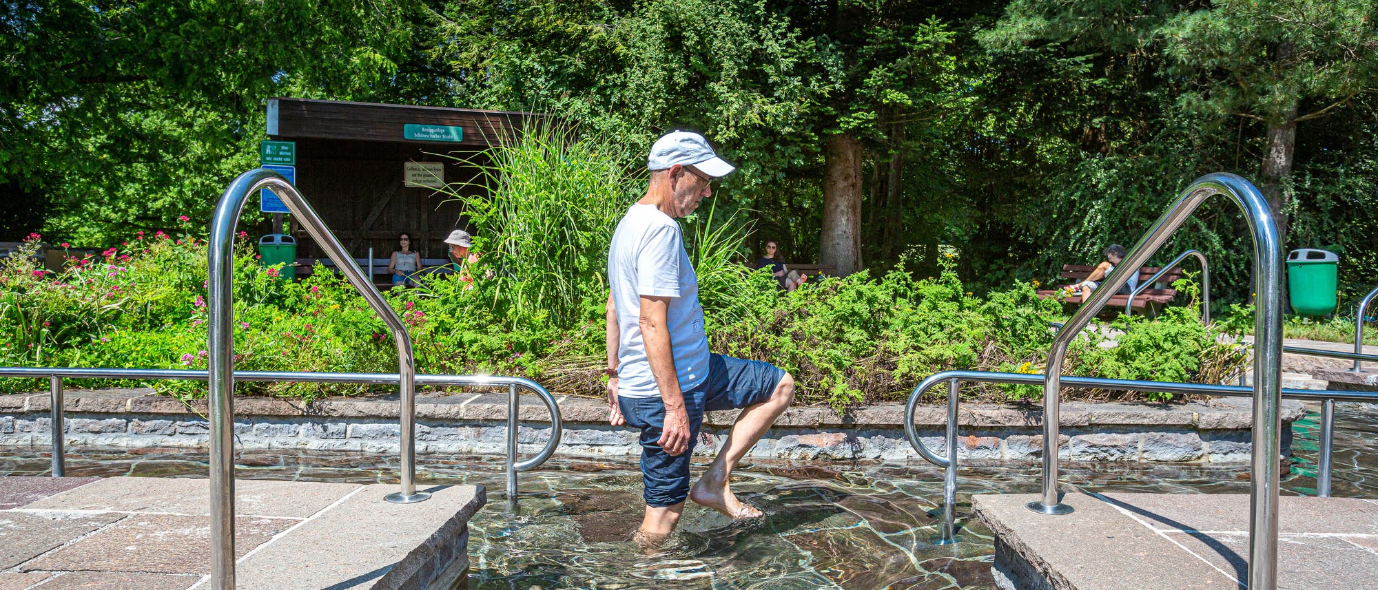 Eine Person geht durch ein Kneippbecken im Freien, umgeben von üppigem Grün und Bäumen. Der Himmel ist klar, und die Sonne scheint. Im Hintergrund sind weitere Menschen auf Bänken zu sehen. Edelstahlgeländer flankieren den Weg durch das Wasser.