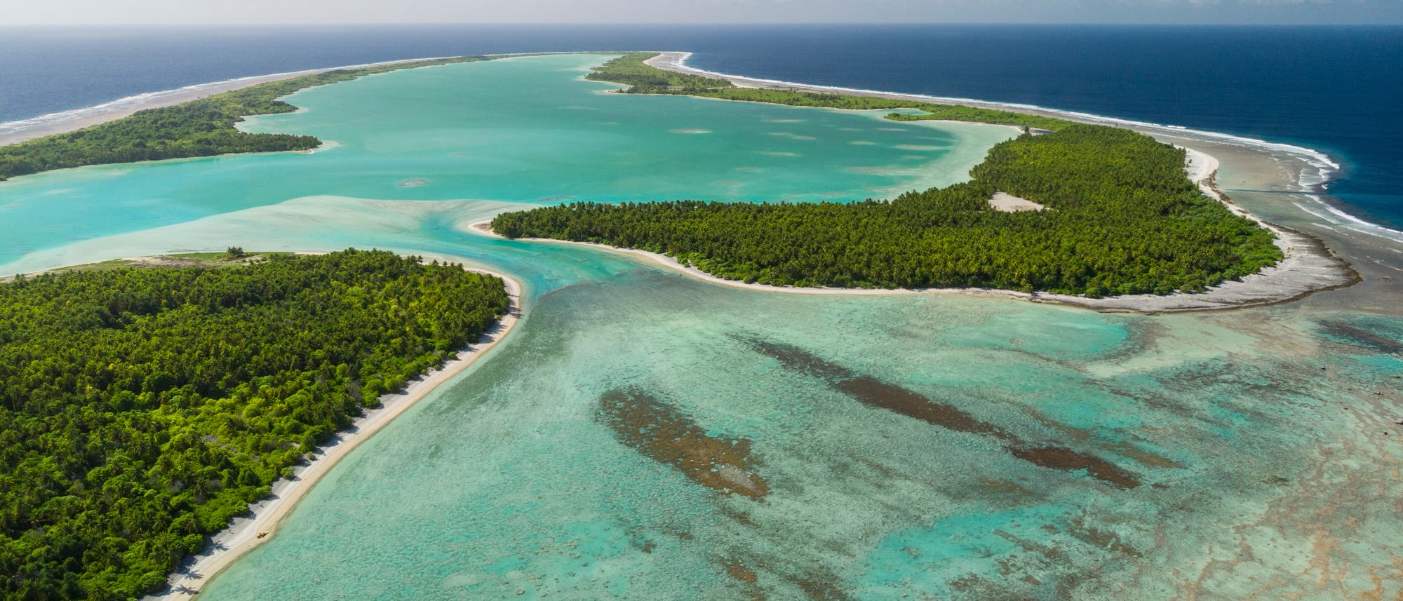 Luftaufnahme einer tropischen Insel mit üppigem, grünem Wald, umgeben von türkisfarbenem Wasser. Die Küstenlinie ist von weißen Sandstränden gesäumt, und das Meer wechselt von hellen zu dunkleren Blautönen. Im Hintergrund erstreckt sich der Ozean bis zum Horizont unter einem klaren, blauen Himmel. Luftaufnahme einer tropischen Insel mit üppigem, grünem Wald, umgeben von türkisfarbenem Wasser. Die Küstenlinie ist von weißen Sandstränden gesäumt, und das Meer wechselt von hellen zu dunkleren Blautönen. Im Hintergrund erstreckt sich der Ozean bis zum Horizont unter einem klaren, blauen Himmel.