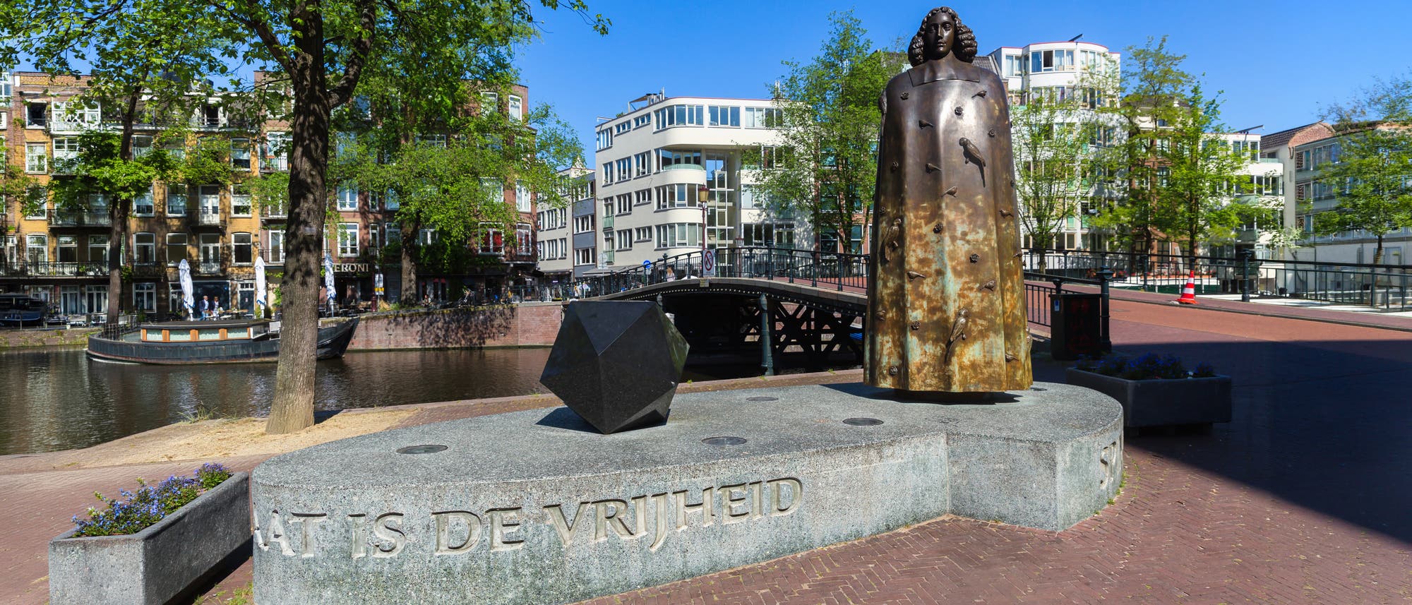 Eine Bronzestatue steht auf einem steinernen Sockel in einer st&auml;dtischen Umgebung neben einem Kanal. Der Sockel tr&auml;gt die Inschrift &bdquo;DAT IS DE VRIJHEID&ldquo;. Im Hintergrund sind moderne und historische Geb&auml;ude sowie eine Br&uuml;cke zu sehen. Ein Baum spendet Schatten, und der Himmel ist klar und blau.
