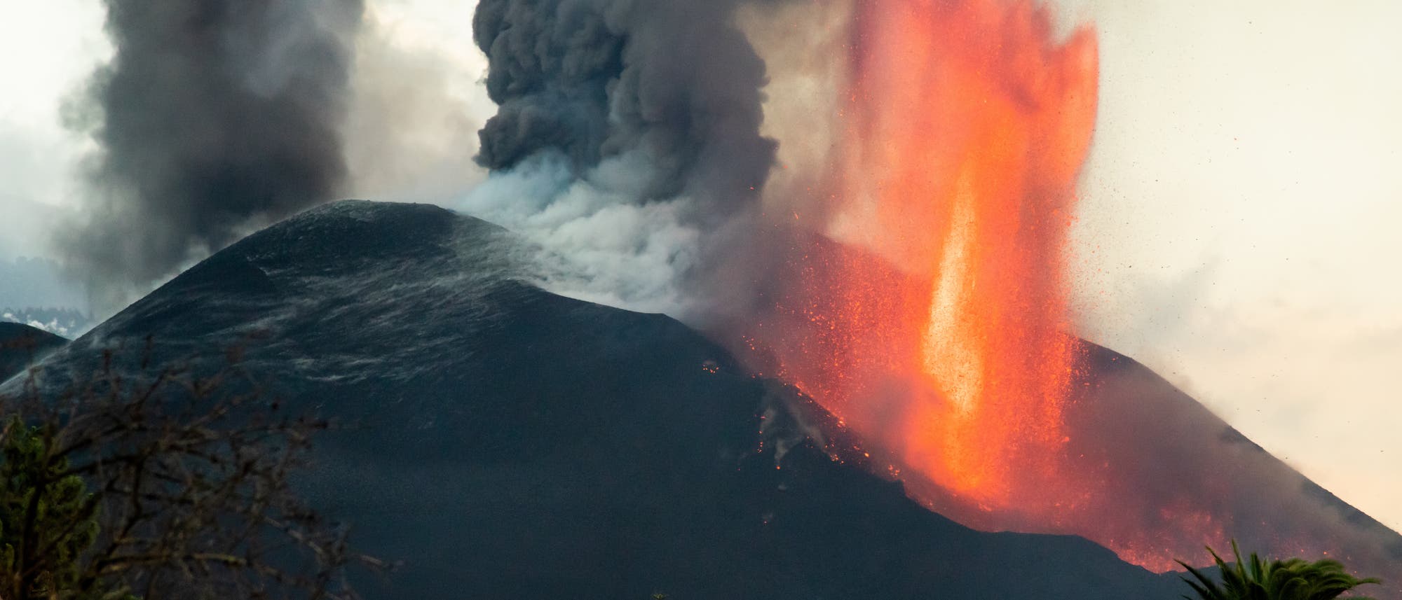 Ein Vulkan bricht aus und stößt eine große Menge glühender Lava und dichten schwarzen Rauch in die Luft. Die Lavafontäne leuchtet hellorange und hebt sich gegen den dunklen Vulkanhügel ab. Im Vordergrund sind einige Bäume zu sehen, die die Szenerie einrahmen. Der Himmel ist teilweise von Rauch verdeckt. Ein Vulkan bricht aus und stößt eine große Menge glühender Lava und dichten schwarzen Rauch in die Luft. Die Lavafontäne leuchtet hellorange und hebt sich gegen den dunklen Vulkanhügel ab. Im Vordergrund sind einige Bäume zu sehen, die die Szenerie einrahmen. Der Himmel ist teilweise von Rauch verdeckt.