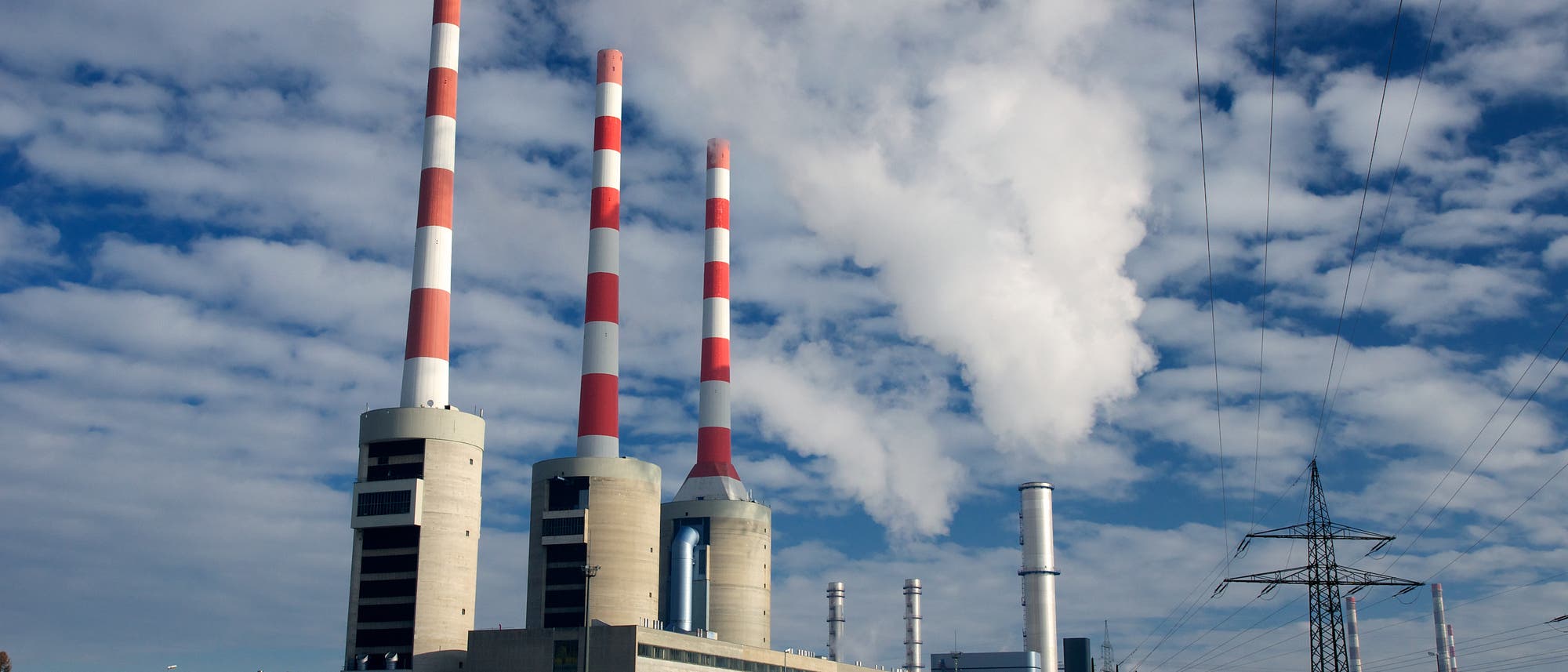 Ein gro&szlig;es Kraftwerk mit mehreren hohen Schornsteinen, die abwechselnd rot-wei&szlig; gestreift sind, ragt in den Himmel. Aus einem der Schornsteine steigt wei&szlig;er Rauch auf. Im Vordergrund sind Stromleitungen und ein verschneites Feld zu sehen. Der Himmel ist teils bew&ouml;lkt.