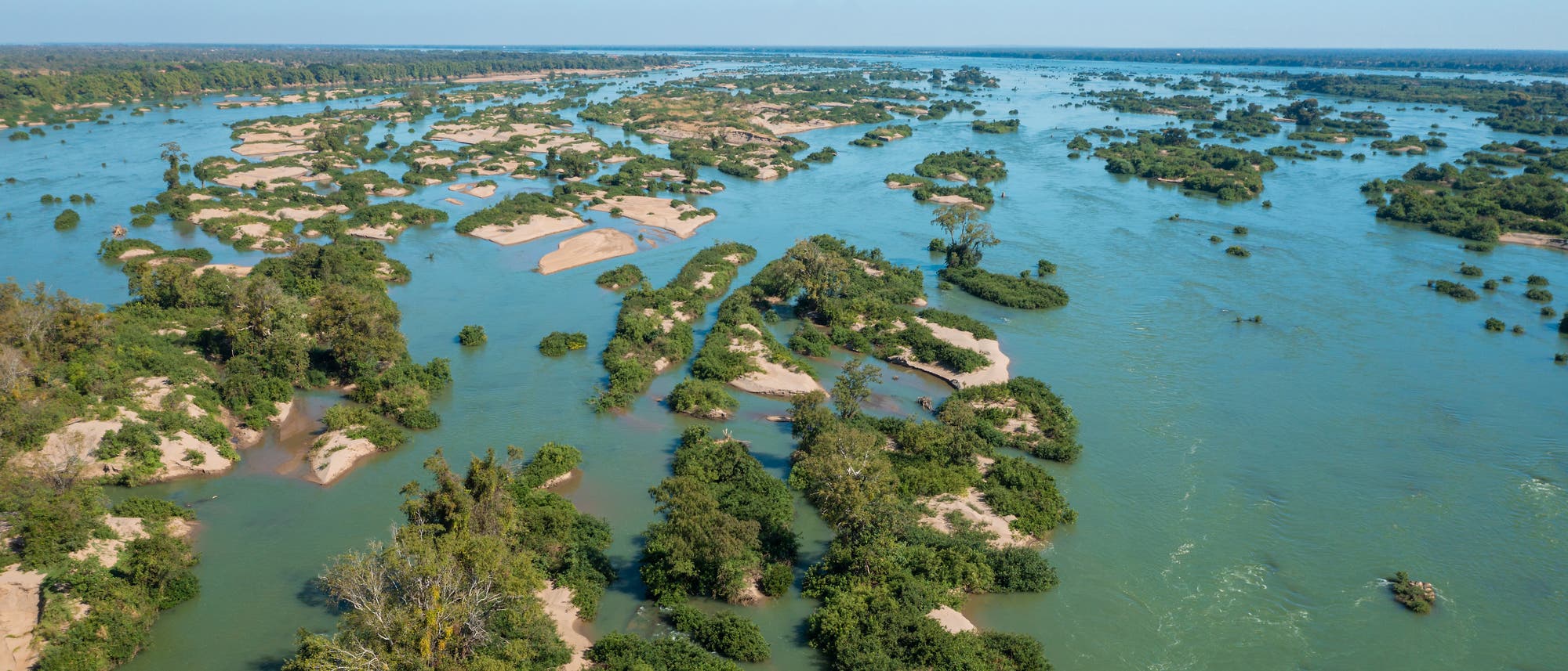 Luftaufnahme eines weitl&auml;ufigen Flussdeltas mit zahlreichen kleinen Inseln, die von &uuml;ppigem Gr&uuml;n bedeckt sind. Das Wasser des Flusses ist blaugr&uuml;n und flie&szlig;t ruhig zwischen den Inseln hindurch. Der Horizont zeigt eine dichte Baumlinie unter einem klaren blauen Himmel.