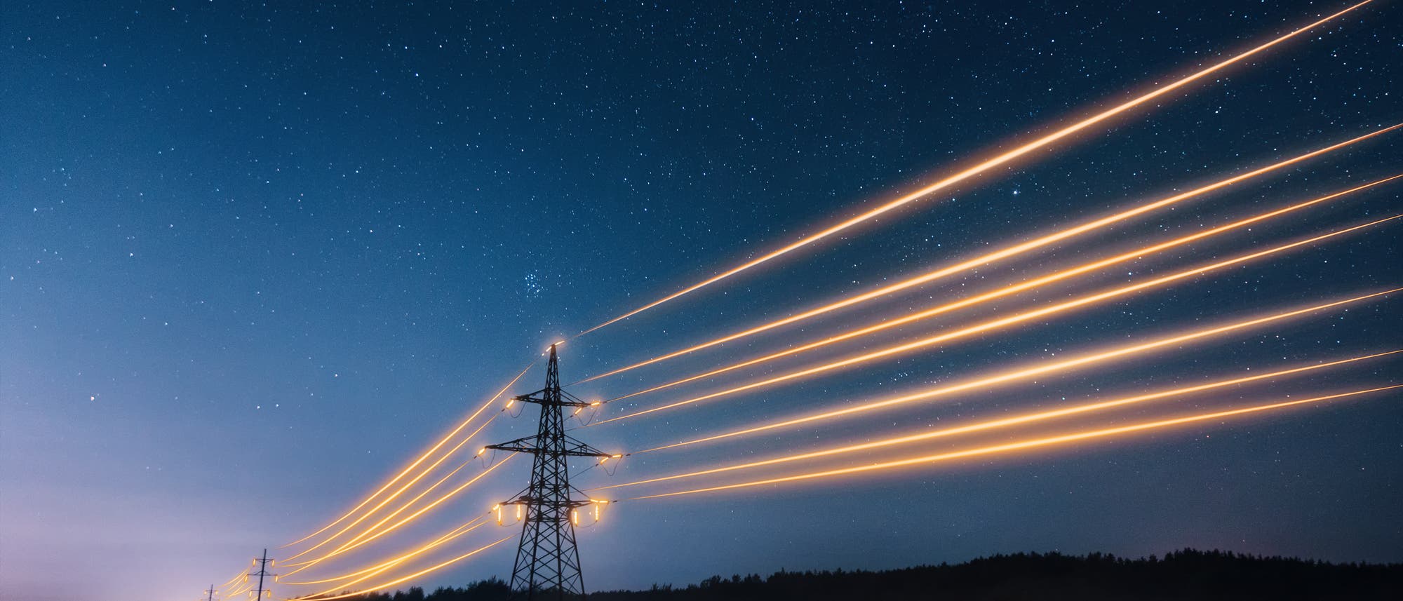 Ein nächtliches Foto zeigt eine Reihe von Strommasten, die sich durch eine Landschaft erstrecken. Die Masten sind mit leuchtenden Stromleitungen verbunden, die in langen, geraden Linien verlaufen. Der Himmel ist klar und voller Sterne, was der Szene eine ruhige und eindrucksvolle Atmosphäre verleiht. Die Lichter der Stromleitungen heben sich kontrastreich vom dunklen Himmel ab.