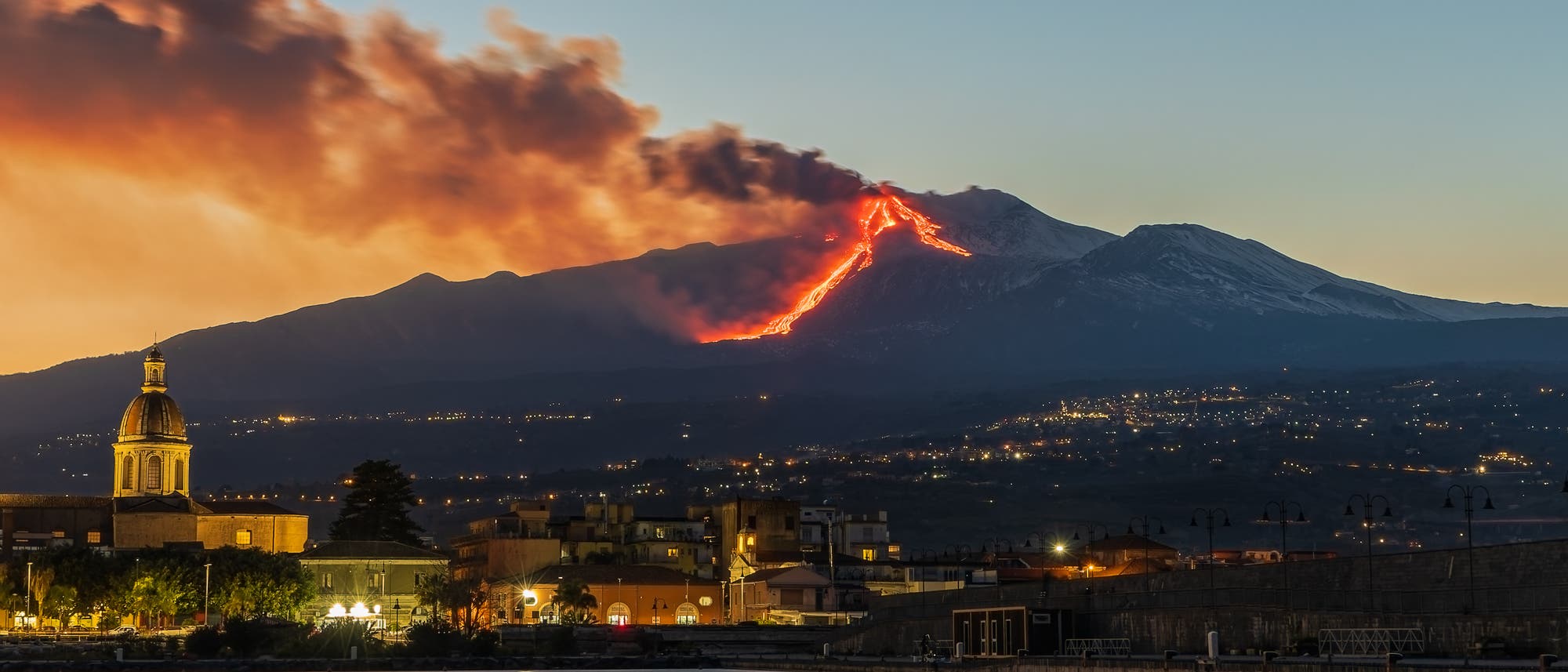 Eine nächtliche Landschaft zeigt einen Vulkan, der Lava ausstößt, während Rauch in den Himmel aufsteigt. Im Vordergrund ist eine Stadt mit beleuchteten Gebäuden und einer Kirche mit Kuppel zu sehen. Der Vulkan leuchtet rot-orange, was einen starken Kontrast zum dunklen Himmel und der ruhigen Wasseroberfläche im Vordergrund bildet. Eine nächtliche Landschaft zeigt einen Vulkan, der Lava ausstößt, während Rauch in den Himmel aufsteigt. Im Vordergrund ist eine Stadt mit beleuchteten Gebäuden und einer Kirche mit Kuppel zu sehen. Der Vulkan leuchtet rot-orange, was einen starken Kontrast zum dunklen Himmel und der ruhigen Wasseroberfläche im Vordergrund bildet.