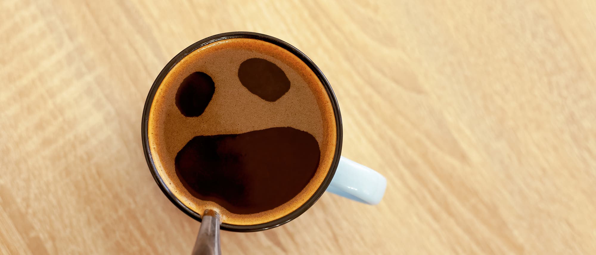 Blick von oben auf eine Kaffeetasse; der Schaum hat drei Öffnungen wie zwei Augen und offener Mund.