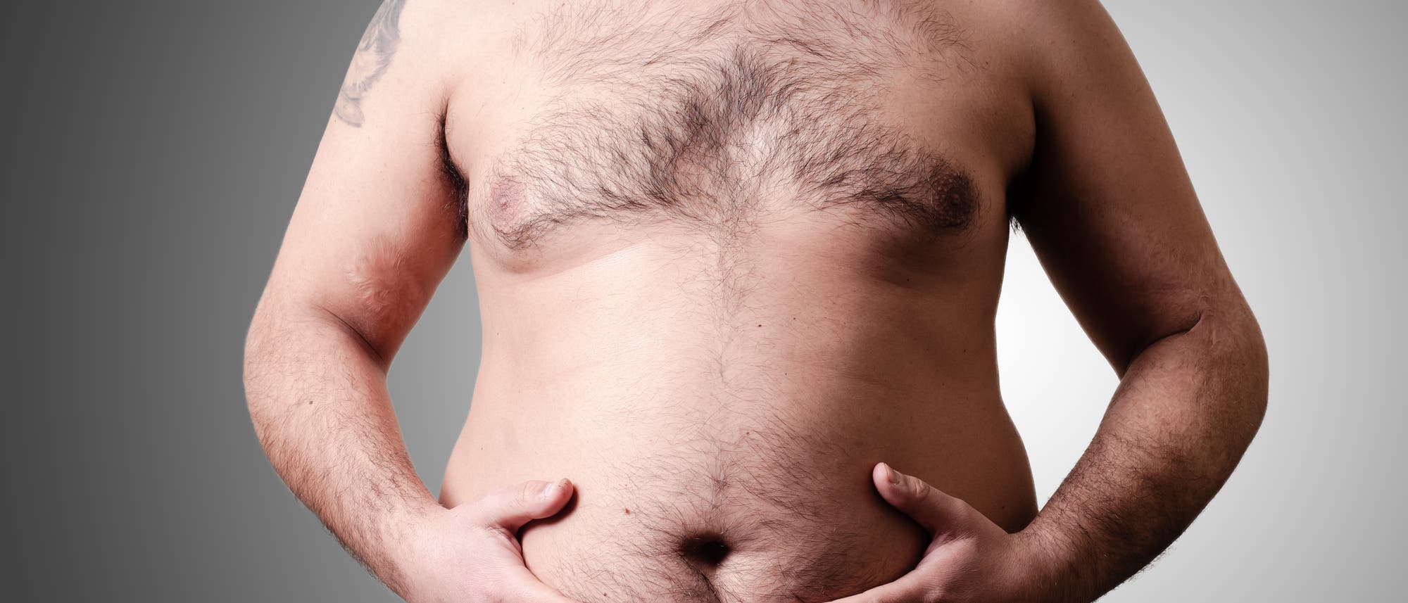 Ein Mann steht vor einem neutralen Hintergrund und h&auml;lt seine H&auml;nde an seinen Bauch. Der Fokus liegt auf dem Oberk&ouml;rper.