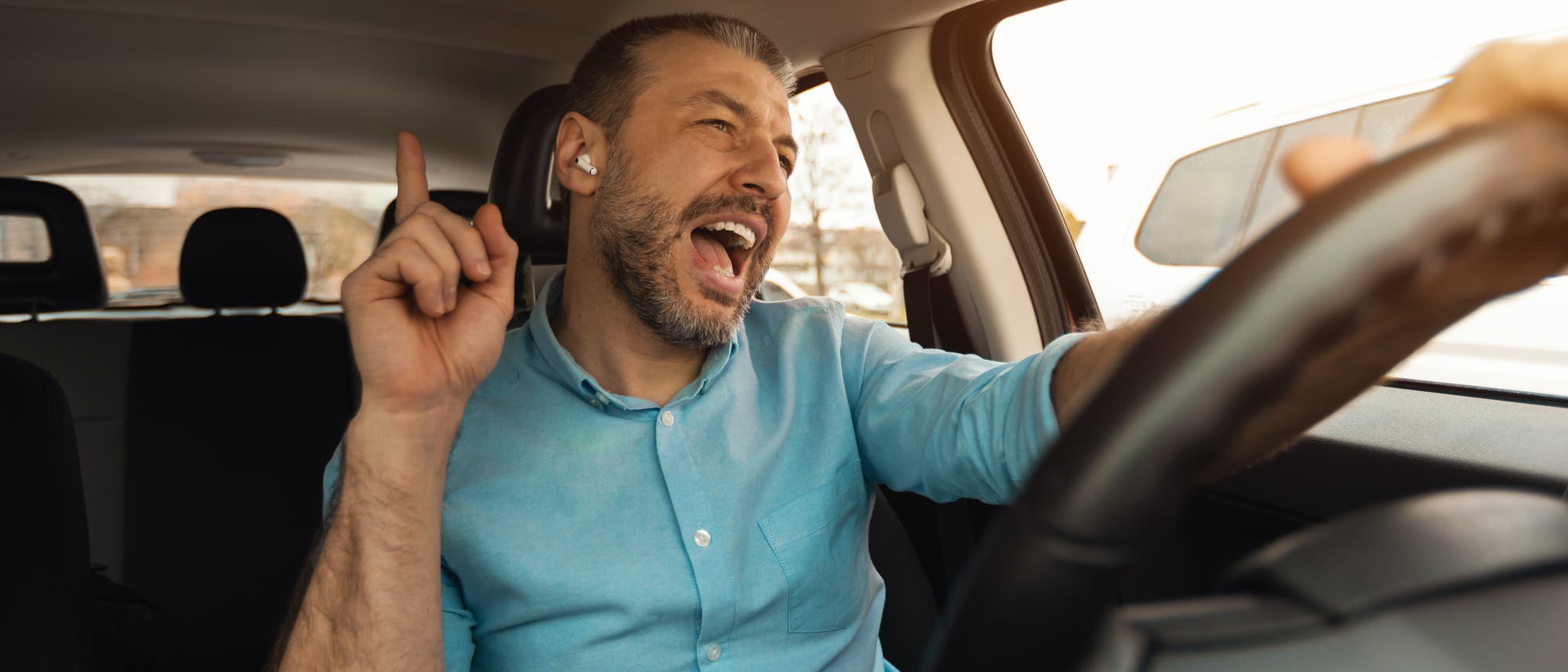 Eine Person sitzt in einem Auto und singt fröhlich, während sie fährt. Sie trägt ein blaues Hemd und hat kabellose Ohrhörer im Ohr. Die rechte Hand hält das Lenkrad, während die linke Hand in die Luft zeigt. Das Bild vermittelt eine entspannte und freudige Stimmung. Eine Person sitzt in einem Auto und singt fröhlich, während sie fährt. Sie trägt ein blaues Hemd und hat kabellose Ohrhörer im Ohr. Die rechte Hand hält das Lenkrad, während die linke Hand in die Luft zeigt. Das Bild vermittelt eine entspannte und freudige Stimmung.
