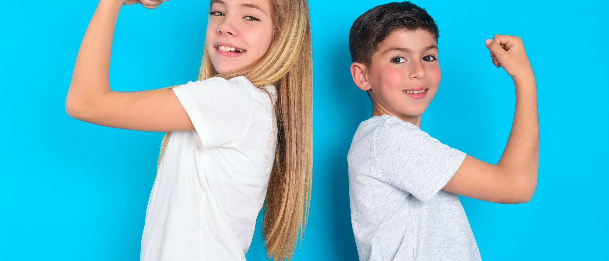 Zwei Kinder stehen Rücken an Rücken vor einem blauen Hintergrund. Beide tragen weiße T-Shirts und zeigen ihre Muskeln, indem sie die Arme anwinkeln und die Fäuste ballen. Sie lächeln in die Kamera. Zwei Kinder stehen Rücken an Rücken vor einem blauen Hintergrund. Beide tragen weiße T-Shirts und zeigen ihre Muskeln, indem sie die Arme anwinkeln und die Fäuste ballen. Sie lächeln in die Kamera.