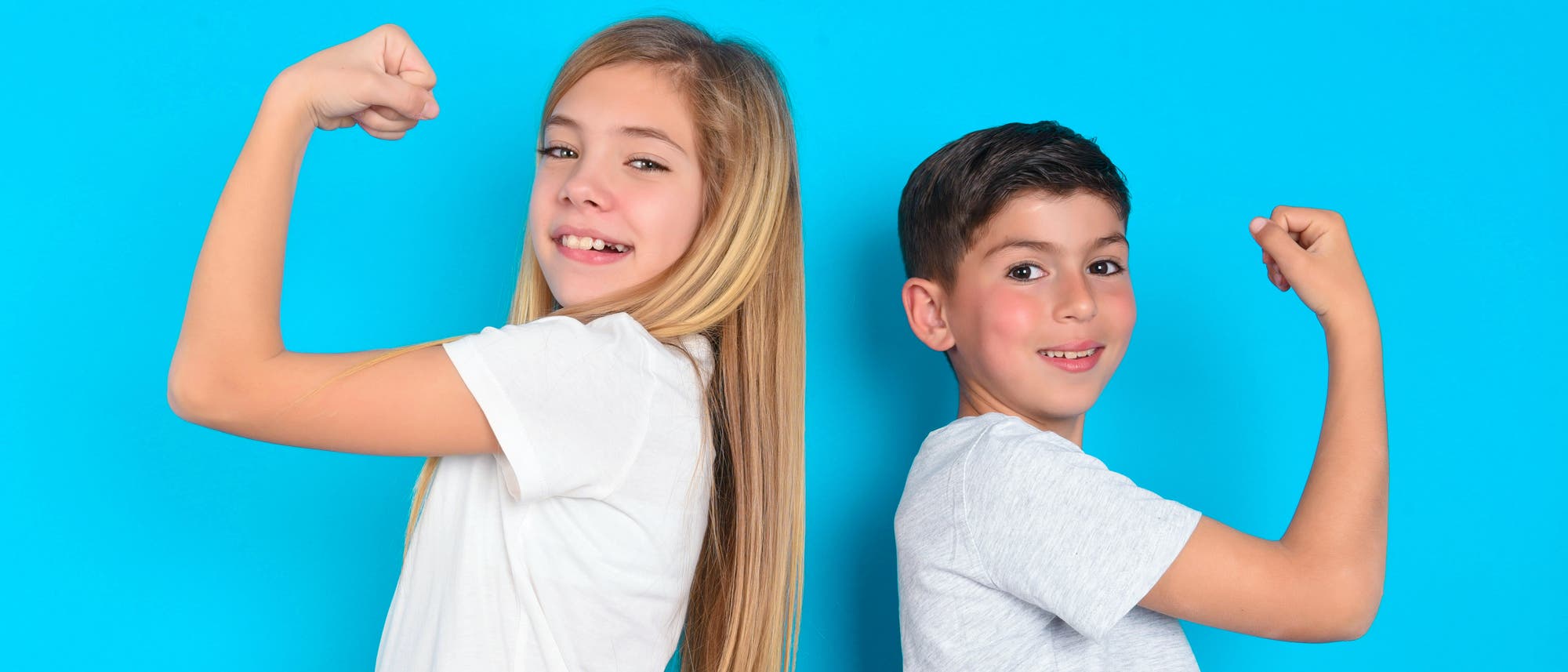 Zwei Kinder stehen R&uuml;cken an R&uuml;cken vor einem blauen Hintergrund und zeigen ihre Muskeln, indem sie die Arme beugen. Beide tragen wei&szlig;e T-Shirts und l&auml;cheln in die Kamera.