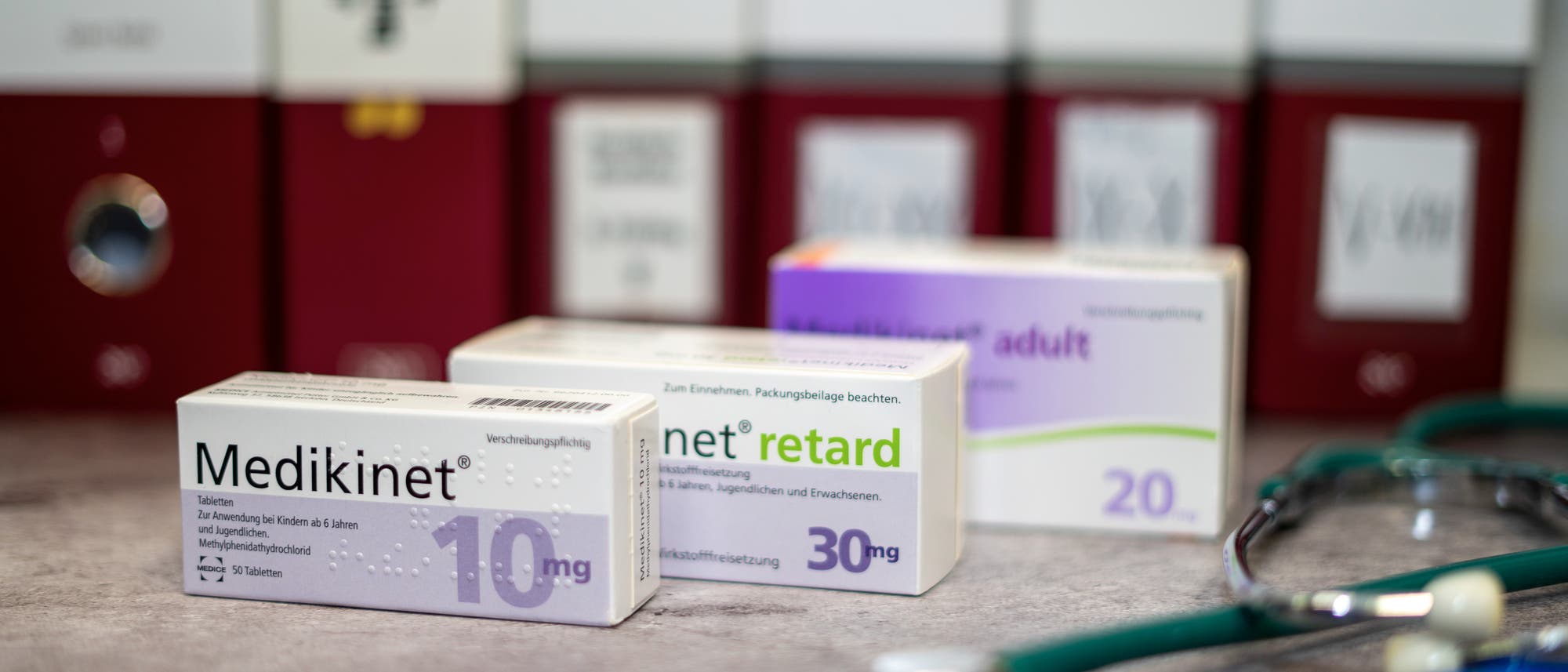 Eine Nahaufnahme von drei Medikamentenschachteln auf einem Tisch, umgeben von roten Aktenordnern im Hintergrund. Die Schachteln sind mit den Aufschriften „Medikinet 10 mg“, „Medikinet retard 30 mg“ und „Medikinet adult 20 mg“ versehen. Ein Stethoskop liegt rechts im Vordergrund. Die Szene vermittelt eine medizinische oder pharmazeutische Umgebung. Eine Nahaufnahme von drei Medikamentenschachteln auf einem Tisch, umgeben von roten Aktenordnern im Hintergrund. Die Schachteln sind mit den Aufschriften „Medikinet 10 mg“, „Medikinet retard 30 mg“ und „Medikinet adult 20 mg“ versehen. Ein Stethoskop liegt rechts im Vordergrund. Die Szene vermittelt eine medizinische oder pharmazeutische Umgebung.