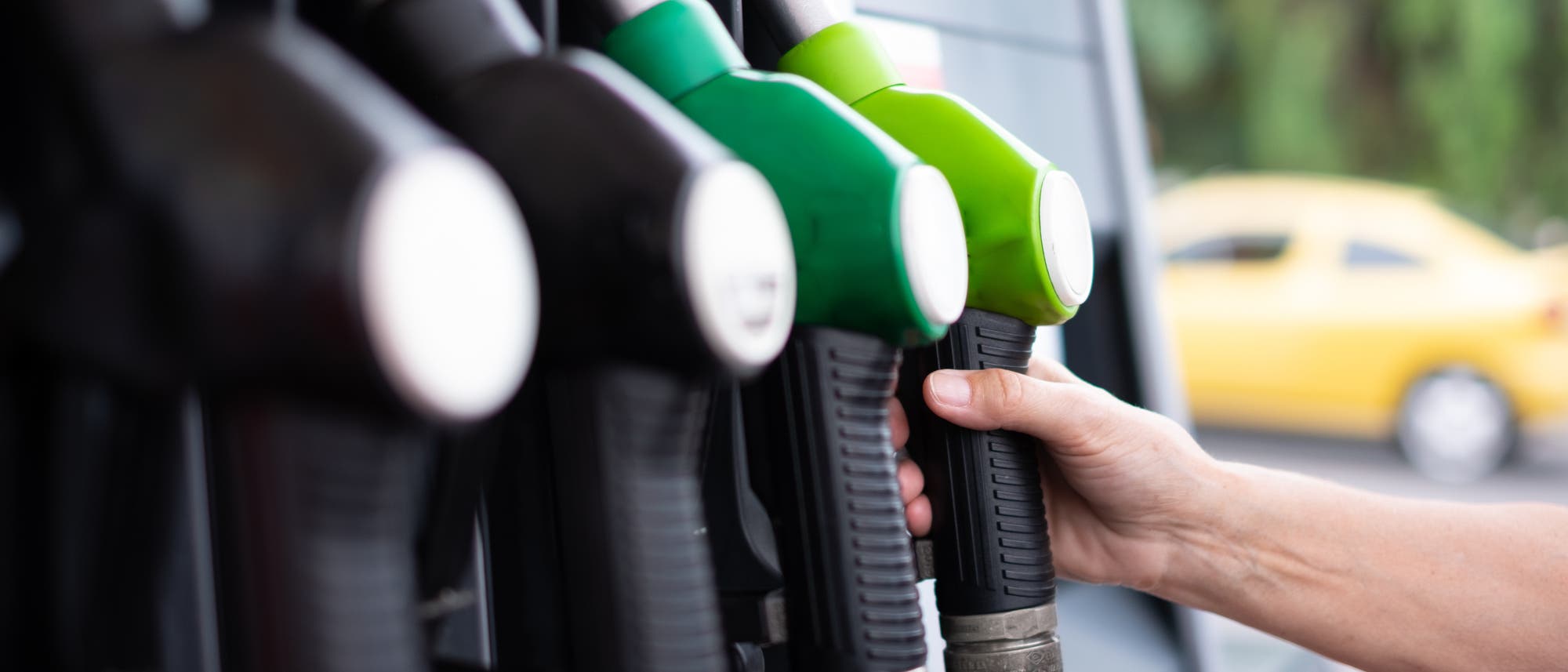Eine Hand greift nach einer gr&uuml;nen Zapfpistole an einer Tankstelle. Mehrere Zapfpistolen sind in einer Reihe angeordnet, darunter schwarze und gr&uuml;ne. Im Hintergrund ist ein gelbes Auto unscharf zu erkennen.