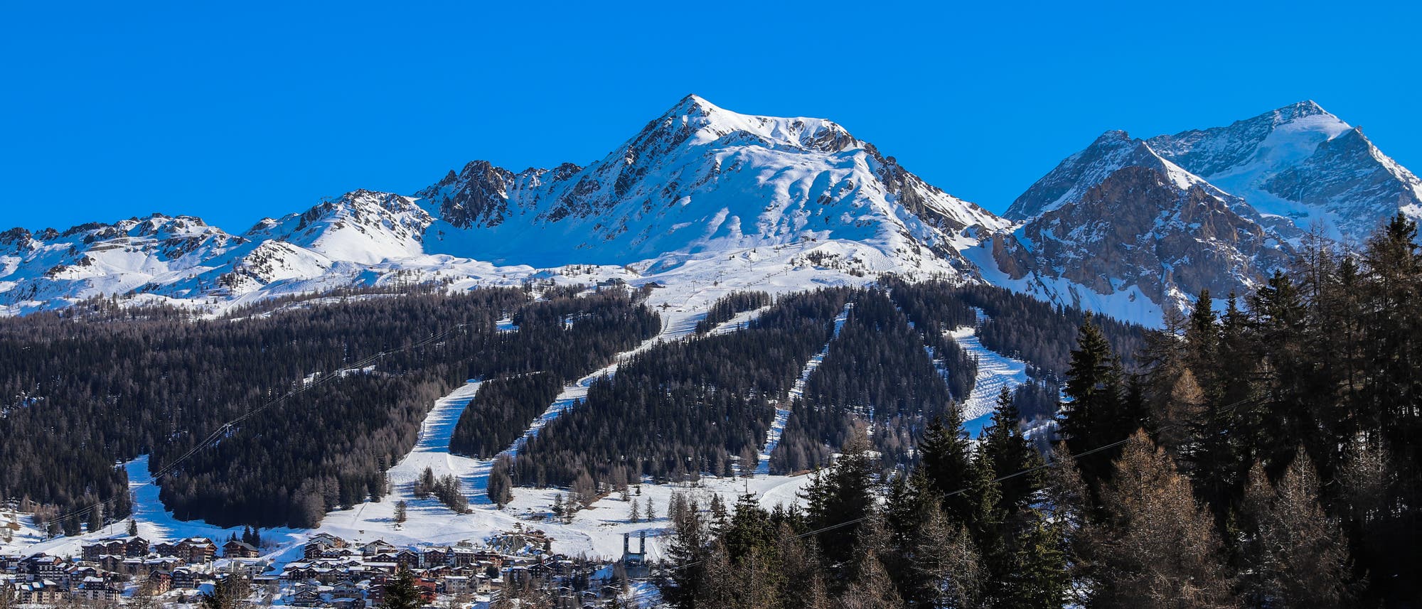 Blick auf einen schneebedeckten Berggipfel unter blauem Himmel. Darunter erstreckt sich ein kleines Dorf mit einigen Dutzend Häusern im Chalet-Stil. Blick auf einen schneebedeckten Berggipfel unter blauem Himmel. Darunter erstreckt sich ein kleines Dorf mit einigen Dutzend Häusern im Chalet-Stil.