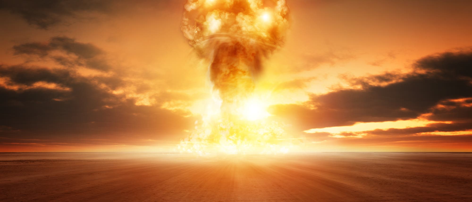 Eine große Explosion in einer Wüstenlandschaft bei Sonnenuntergang. Ein leuchtender Feuerball erhebt sich in die Luft, umgeben von Rauch und Wolken, die den Himmel in warme Orange- und Rottönen färben. Die Szene vermittelt eine dramatische und intensive Atmosphäre.