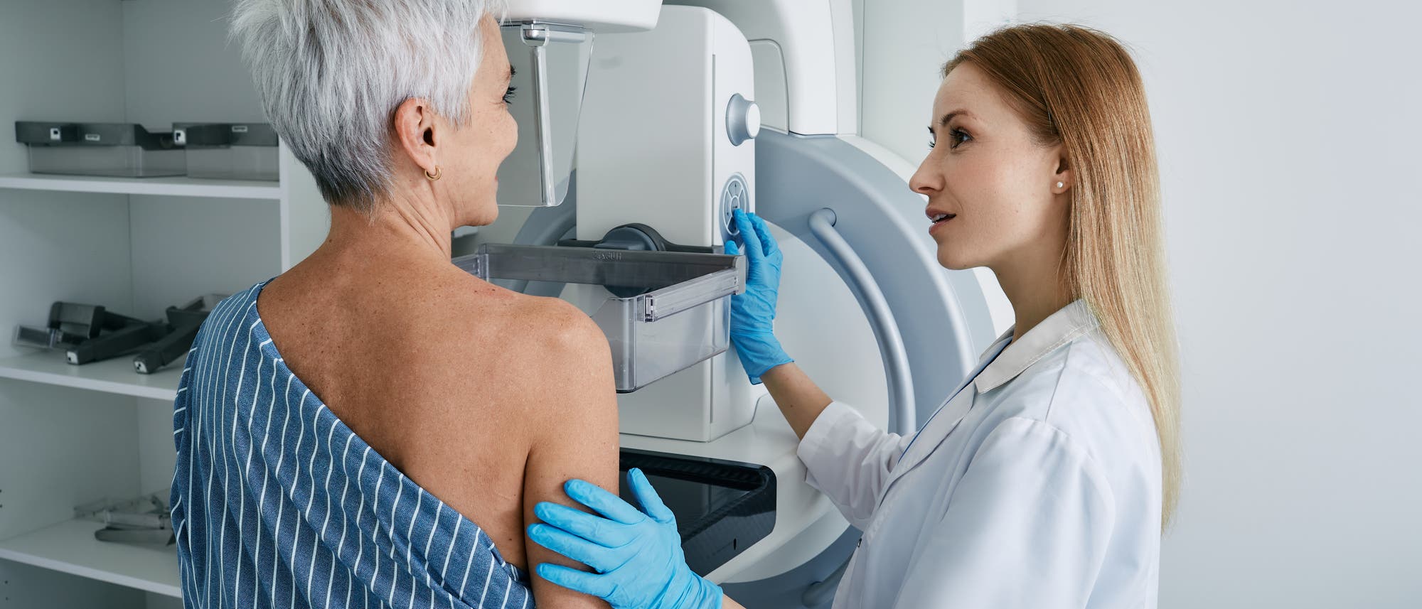 Eine Ärztin in einem weißen Kittel und blauen Handschuhen führt eine Mammographie bei einer Patientin durch. Die Patientin steht vor einem medizinischen Gerät und trägt ein gestreiftes Tuch über einer Schulter. Die Ärztin erklärt den Vorgang und berührt sanft den Arm der Patientin, um sie zu unterstützen. Im Hintergrund sind Regale mit medizinischen Utensilien zu sehen. Eine Ärztin in einem weißen Kittel und blauen Handschuhen führt eine Mammographie bei einer Patientin durch. Die Patientin steht vor einem medizinischen Gerät und trägt ein gestreiftes Tuch über einer Schulter. Die Ärztin erklärt den Vorgang und berührt sanft den Arm der Patientin, um sie zu unterstützen. Im Hintergrund sind Regale mit medizinischen Utensilien zu sehen.