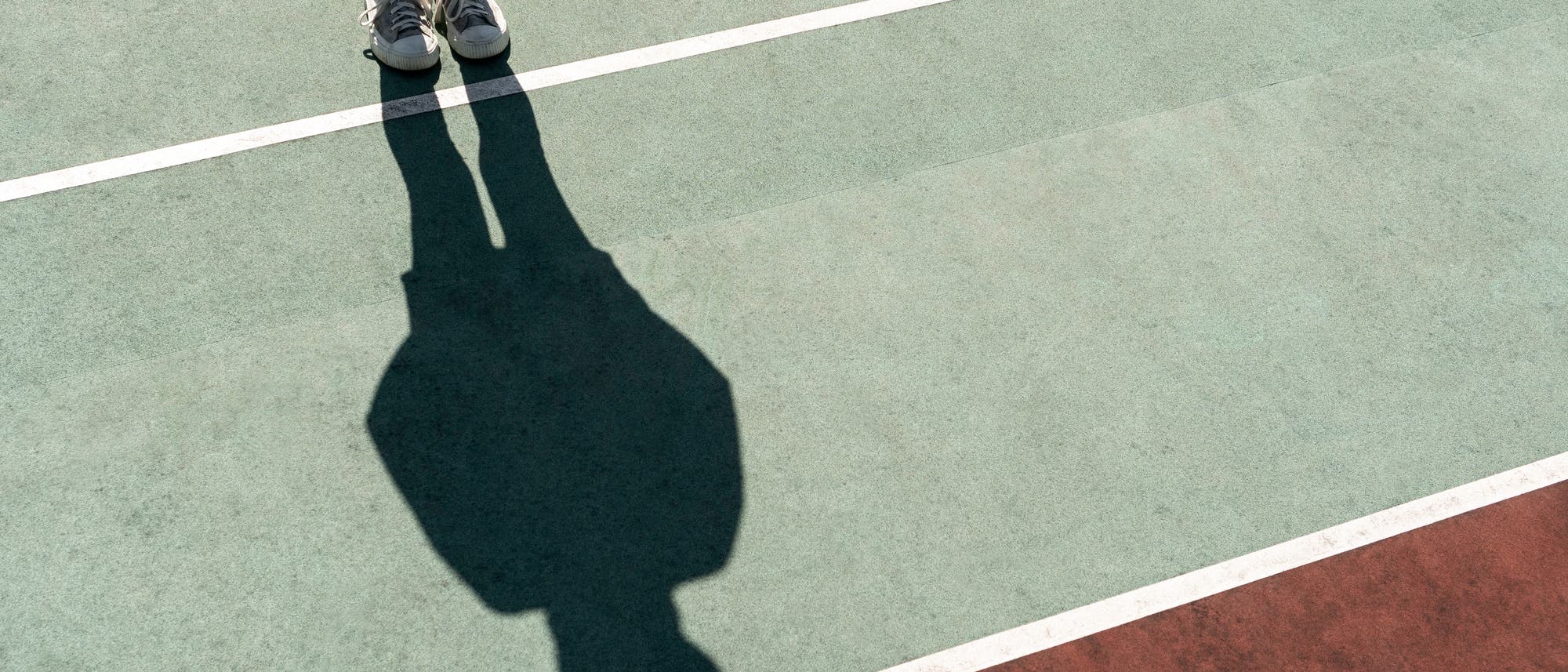 Eine Person steht auf einem Sportplatz mit grünem und rotem Bodenbelag, wobei nur die Beine und Schuhe sichtbar sind. Die Person wirft einen langen Schatten auf den Boden, der von weißen Linien markiert ist. Die Szene ist bei hellem Sonnenlicht aufgenommen. Eine Person steht auf einem Sportplatz mit grünem und rotem Bodenbelag, wobei nur die Beine und Schuhe sichtbar sind. Die Person wirft einen langen Schatten auf den Boden, der von weißen Linien markiert ist. Die Szene ist bei hellem Sonnenlicht aufgenommen.