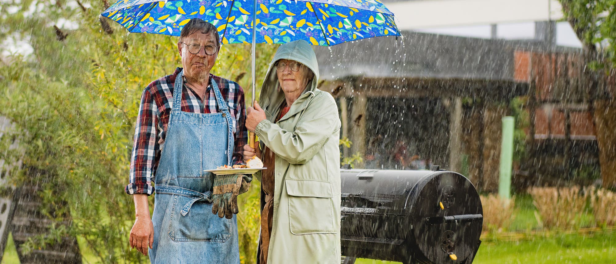 Ein älteres Paar steht im Regen unter einem bunten Regenschirm. Der Mann trägt eine Schürze und hält einen Teller mit Essen, während die Frau einen Regenmantel trägt. Im Hintergrund sind ein Grill und ein Garten zu sehen. Ein älteres Paar steht im Regen unter einem bunten Regenschirm. Der Mann trägt eine Schürze und hält einen Teller mit Essen, während die Frau einen Regenmantel trägt. Im Hintergrund sind ein Grill und ein Garten zu sehen.