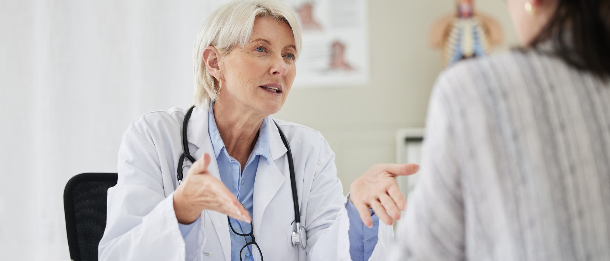 Eine Ärztin mit grauem Haar und Stethoskop spricht mit einer Patientin in einem Behandlungszimmer. Die Ärztin gestikuliert, während sie der Patientin zuhört. Im Hintergrund sind medizinische Diagramme sichtbar. Eine Ärztin mit grauem Haar und Stethoskop spricht mit einer Patientin in einem Behandlungszimmer. Die Ärztin gestikuliert, während sie der Patientin zuhört. Im Hintergrund sind medizinische Diagramme sichtbar.