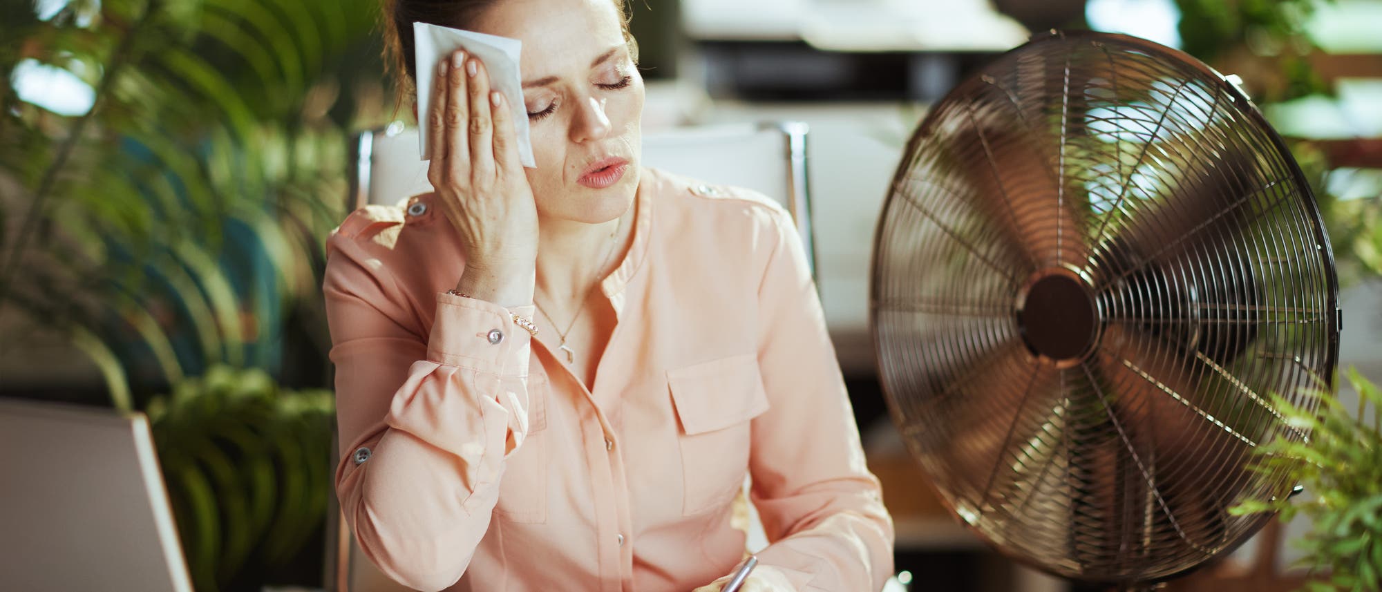 Eine Frau sitzt neben einem Computer und einem Ventilator und hält sich erschöpft ein Tuch an die Stirn