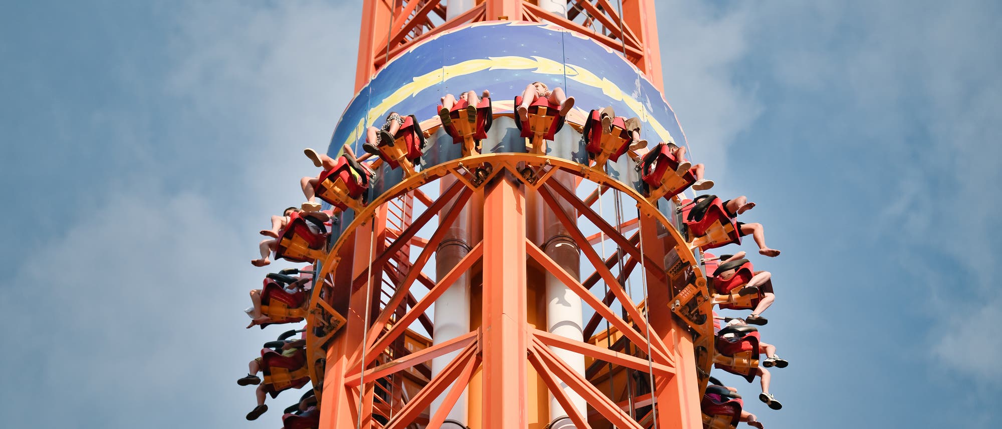 Eine Gruppe von Menschen sitzt in einem kreisförmigen Fahrgeschäft, das an einem hohen, orangefarbenen Turm befestigt ist. Die Personen sind in Sitzen mit Sicherheitsgurten gesichert und scheinen sich in einer schnellen Aufwärtsbewegung zu befinden. Der Himmel ist klar und blau im Hintergrund. Das Fahrgeschäft hat eine bunte Verkleidung mit einem wellenförmigen Muster. Eine Gruppe von Menschen sitzt in einem kreisförmigen Fahrgeschäft, das an einem hohen, orangefarbenen Turm befestigt ist. Die Personen sind in Sitzen mit Sicherheitsgurten gesichert und scheinen sich in einer schnellen Aufwärtsbewegung zu befinden. Der Himmel ist klar und blau im Hintergrund. Das Fahrgeschäft hat eine bunte Verkleidung mit einem wellenförmigen Muster.