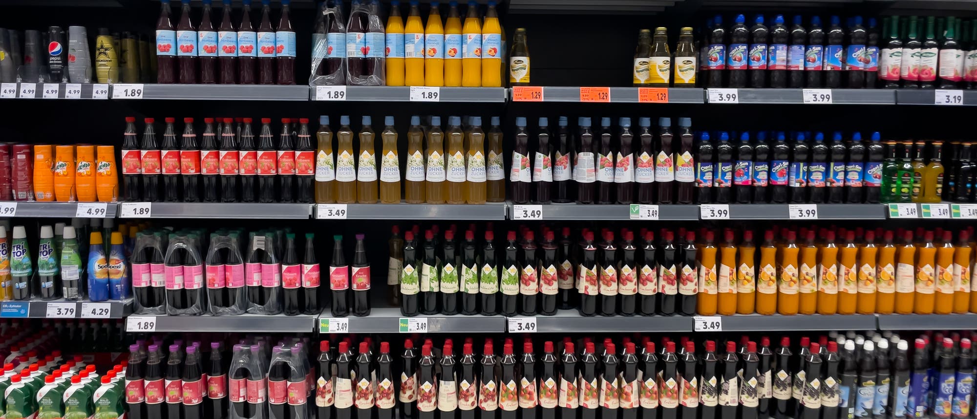 Regale in einem Supermarkt, gef&uuml;llt mit verschiedenen Getr&auml;nkeflaschen. Die Flaschen sind in mehreren Reihen angeordnet und umfassen eine Vielzahl von Marken und Sorten, darunter Softdrinks, S&auml;fte und alkoholische Getr&auml;nke. Preisschilder sind unter den Produkten sichtbar, die Preise reichen von 1,89 &euro; bis 4,99 &euro;. Die Regale sind ordentlich organisiert, und die Flaschen sind in unterschiedlichen Farben und Formen.
