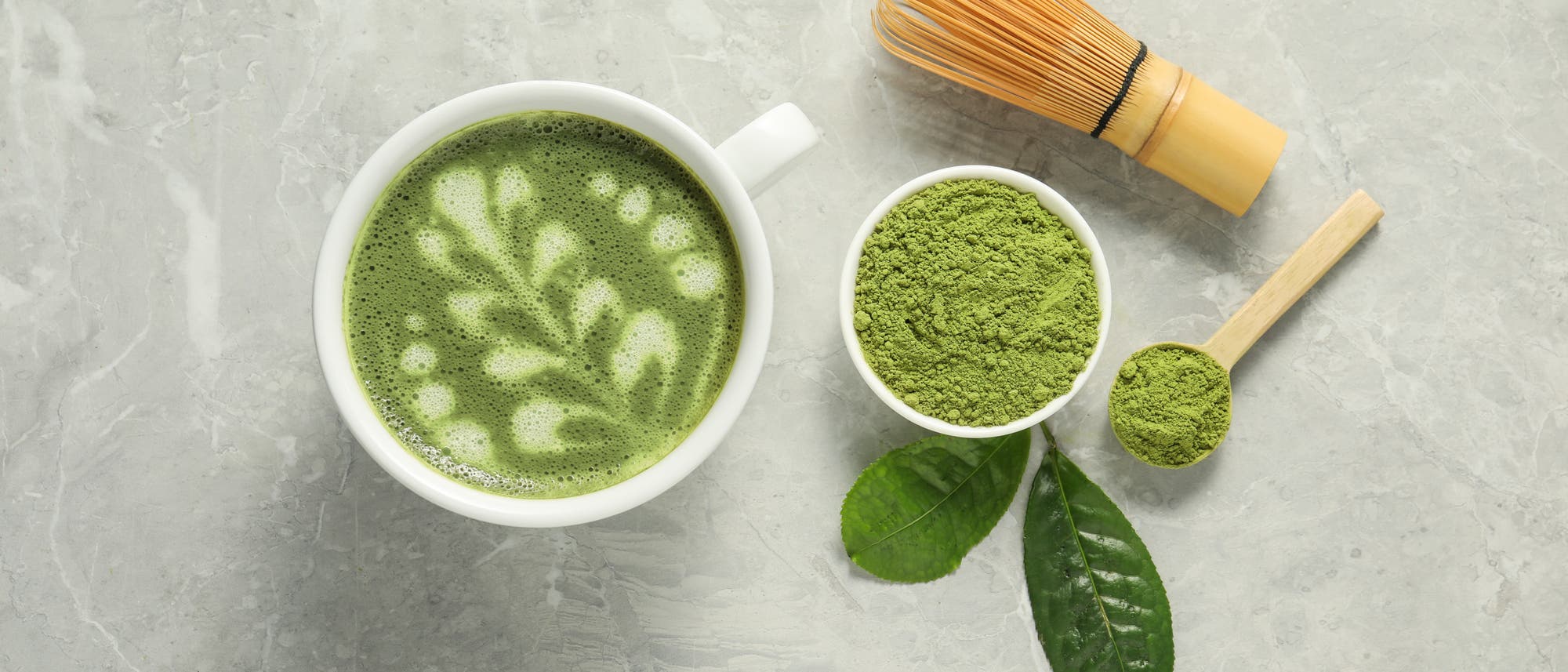 Eine Tasse Matcha-Tee mit einem kunstvollen Blattmuster aus Schaum auf der Oberfl&auml;che. Daneben eine Schale mit gr&uuml;nem Matcha-Pulver, ein Bambusbesen und ein Holzl&ouml;ffel mit Pulver. Zwei gr&uuml;ne Teebl&auml;tter liegen dekorativ auf einer grauen Steinoberfl&auml;che.