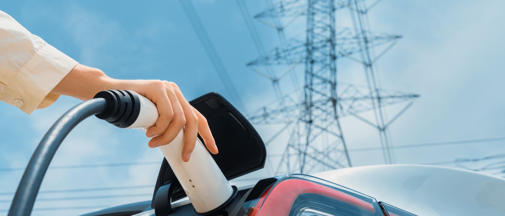 Eine Person lädt ein Elektroauto auf: man sieht ihre Hand, die ein Ladekabel in den Ladeanschluss des Fahrzeugs steckt. Im Hintergrund ist ein Strommast zu sehen, der den Bezug zur Stromversorgung und erneuerbaren Energien herstellt. Der Himmel ist klar und blau.